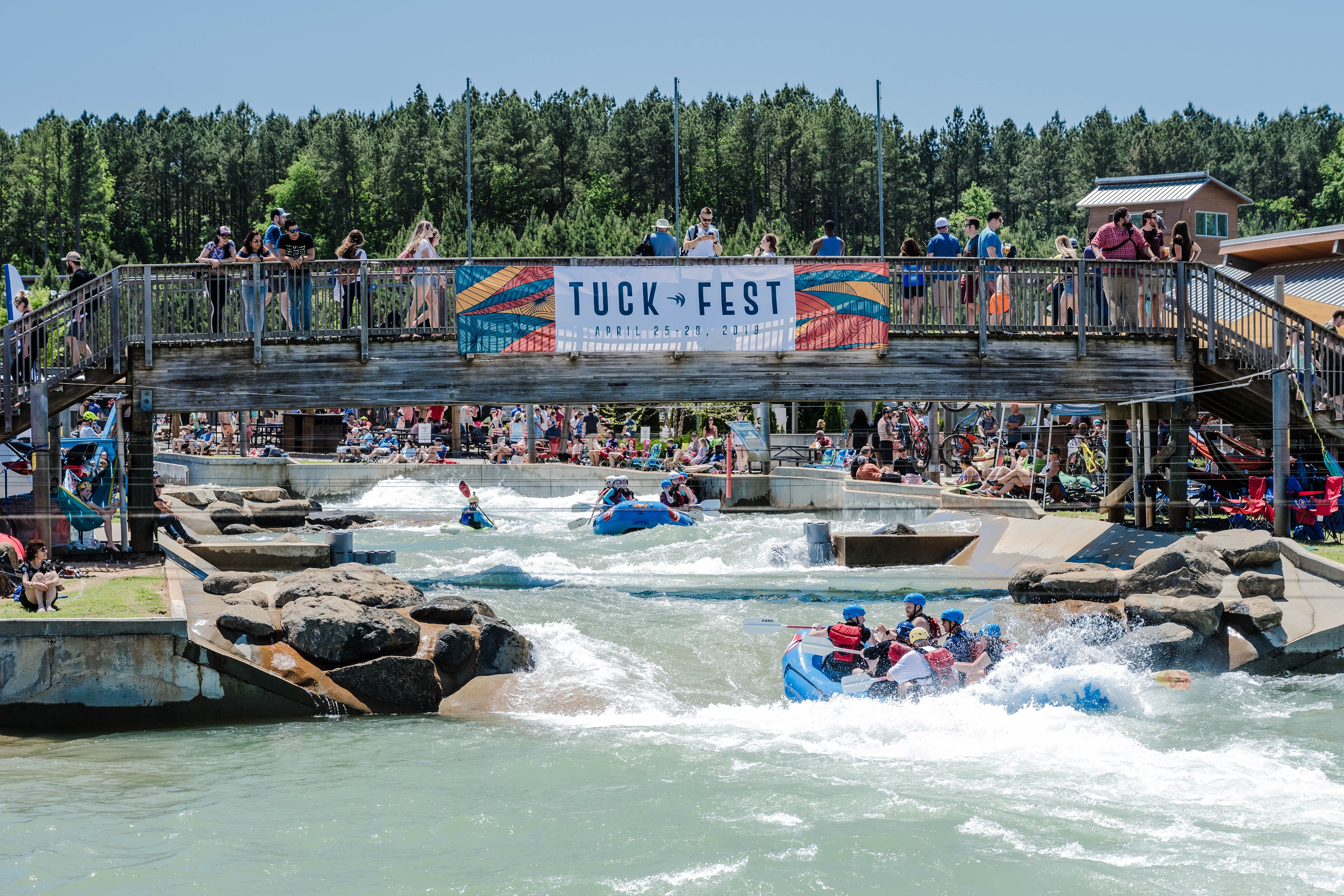 A group of people in a large rubber raft paddle on rushing white water at the US National Whitewater Center in Charlotte, NC. There is a large sign that says