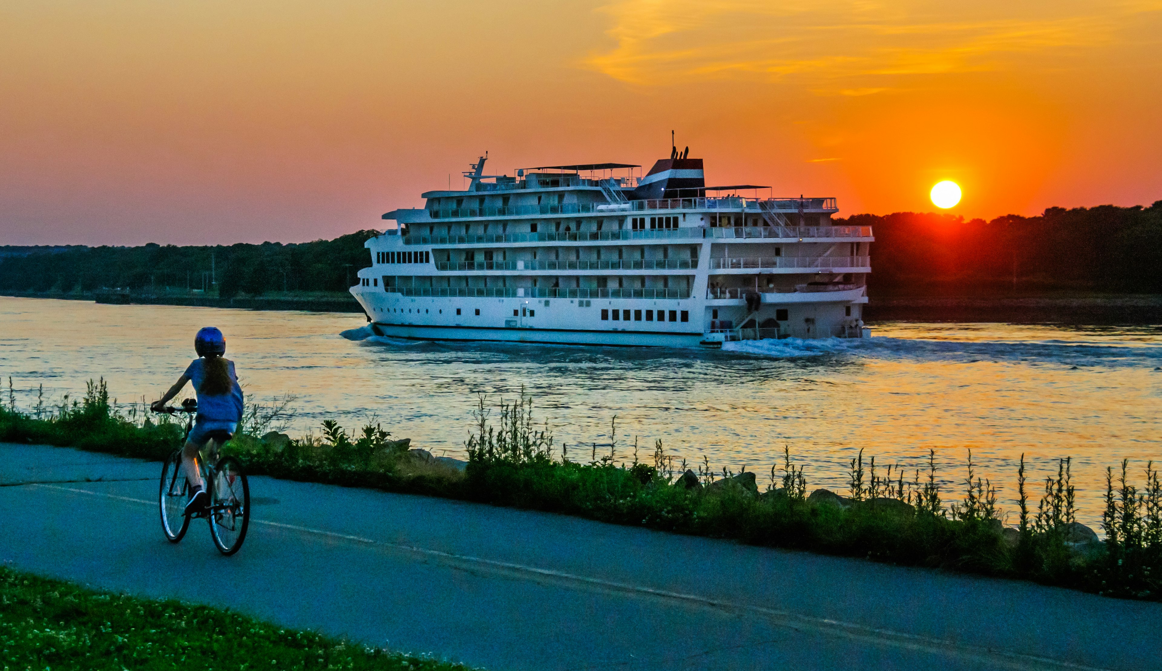 A young woman on a bicycle watches a cruise ship on the Cape Cod Canal