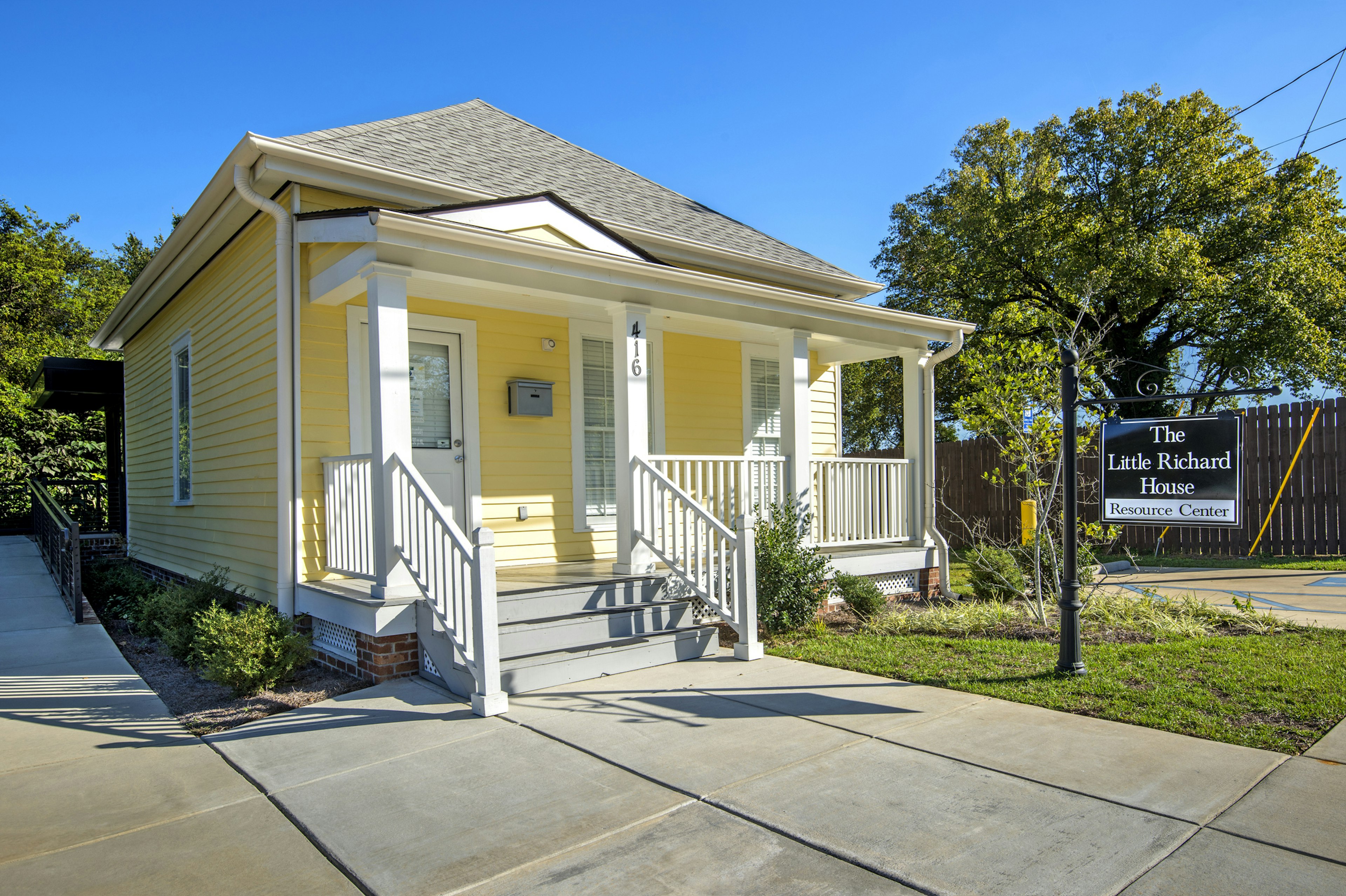 A small wooden house with a veranda painted yellow. A sign hanging outside says The Little Richard House