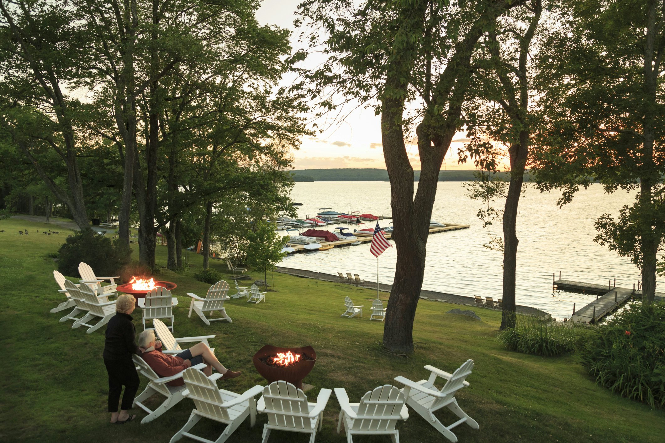 A lake scene with white patio chairs on the grass. A man is seated in one of the chairs with a woman behind his chair him as they gaze out onto the water.
