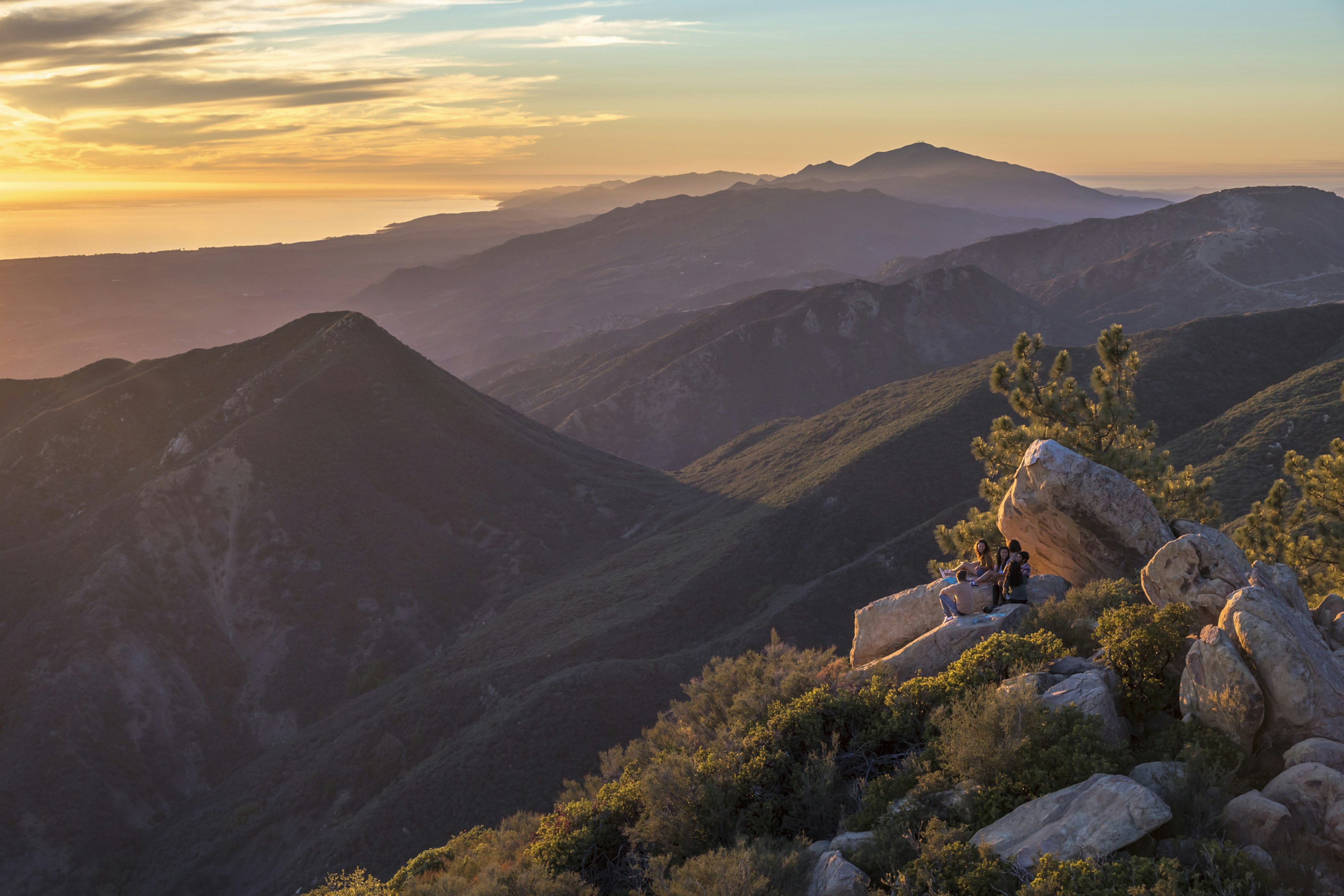 A small group of people sitting on rock on a mountain peak as the sunset casts streaks of orange and yellow across the sky