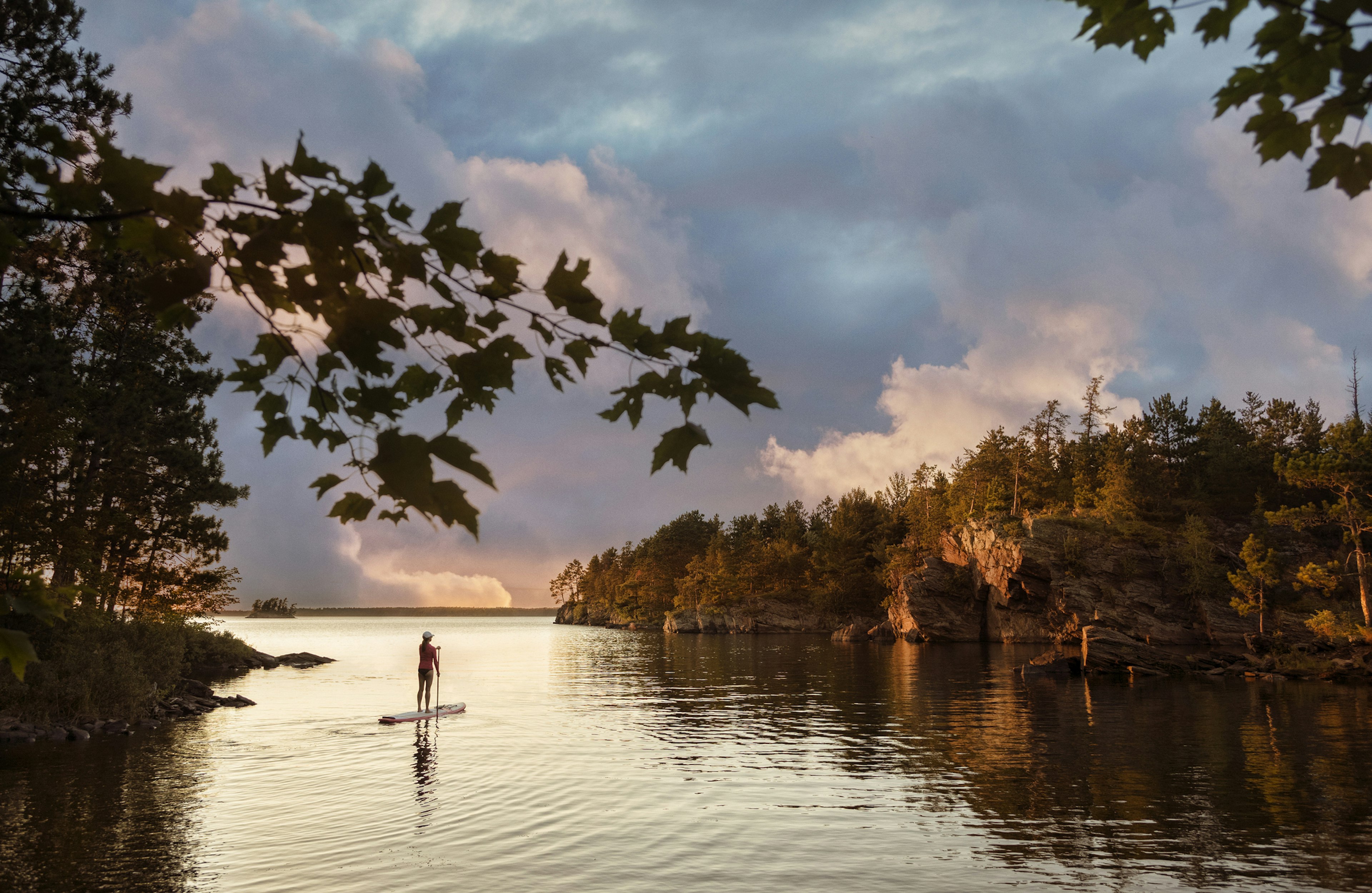 A solo figure on a paddleboard on a lake in a wooded area as the sky turns orange-pink at sunset