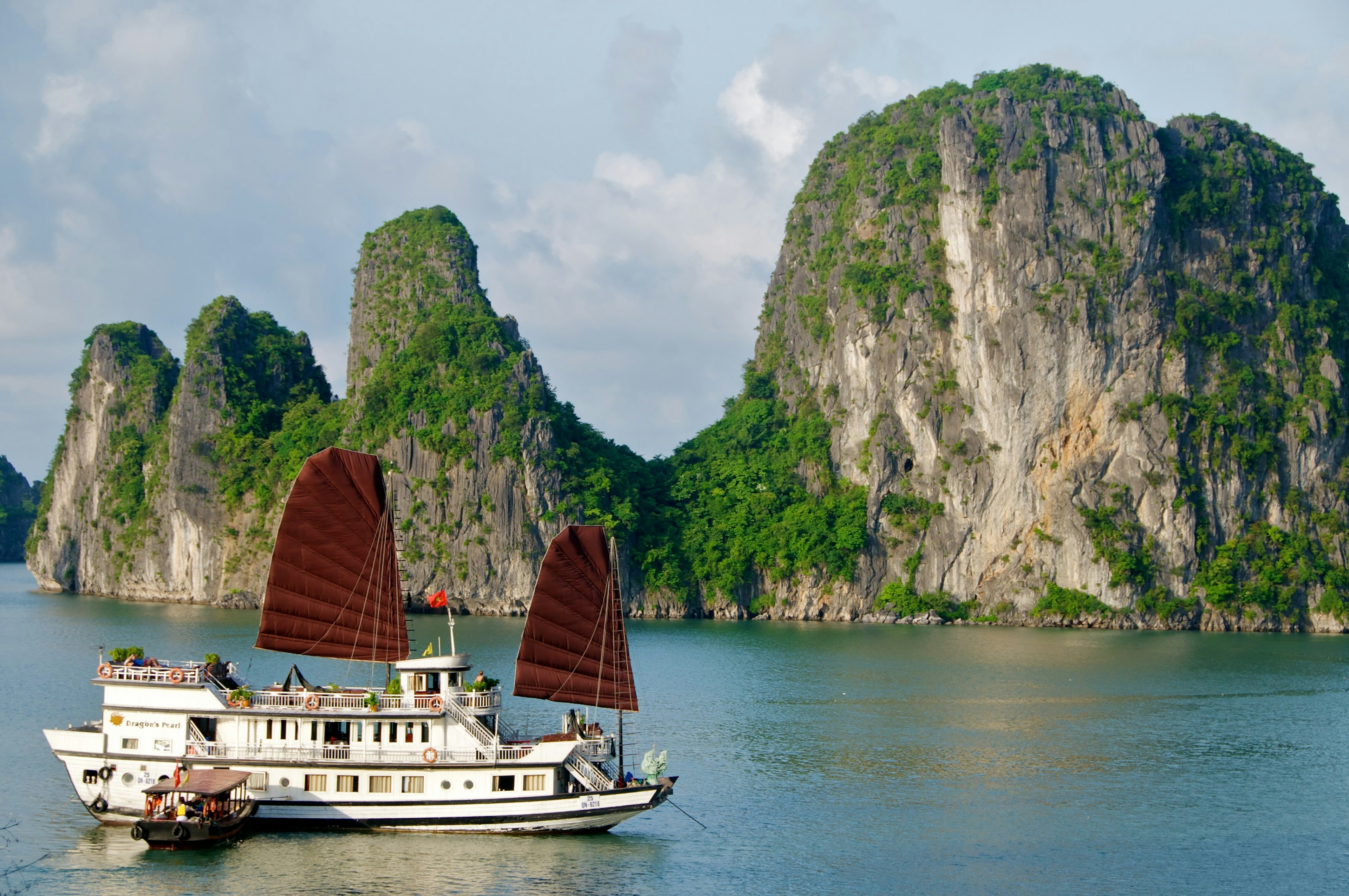 A large boat with red sails is anchored near a huge rocky islet covered in foliage