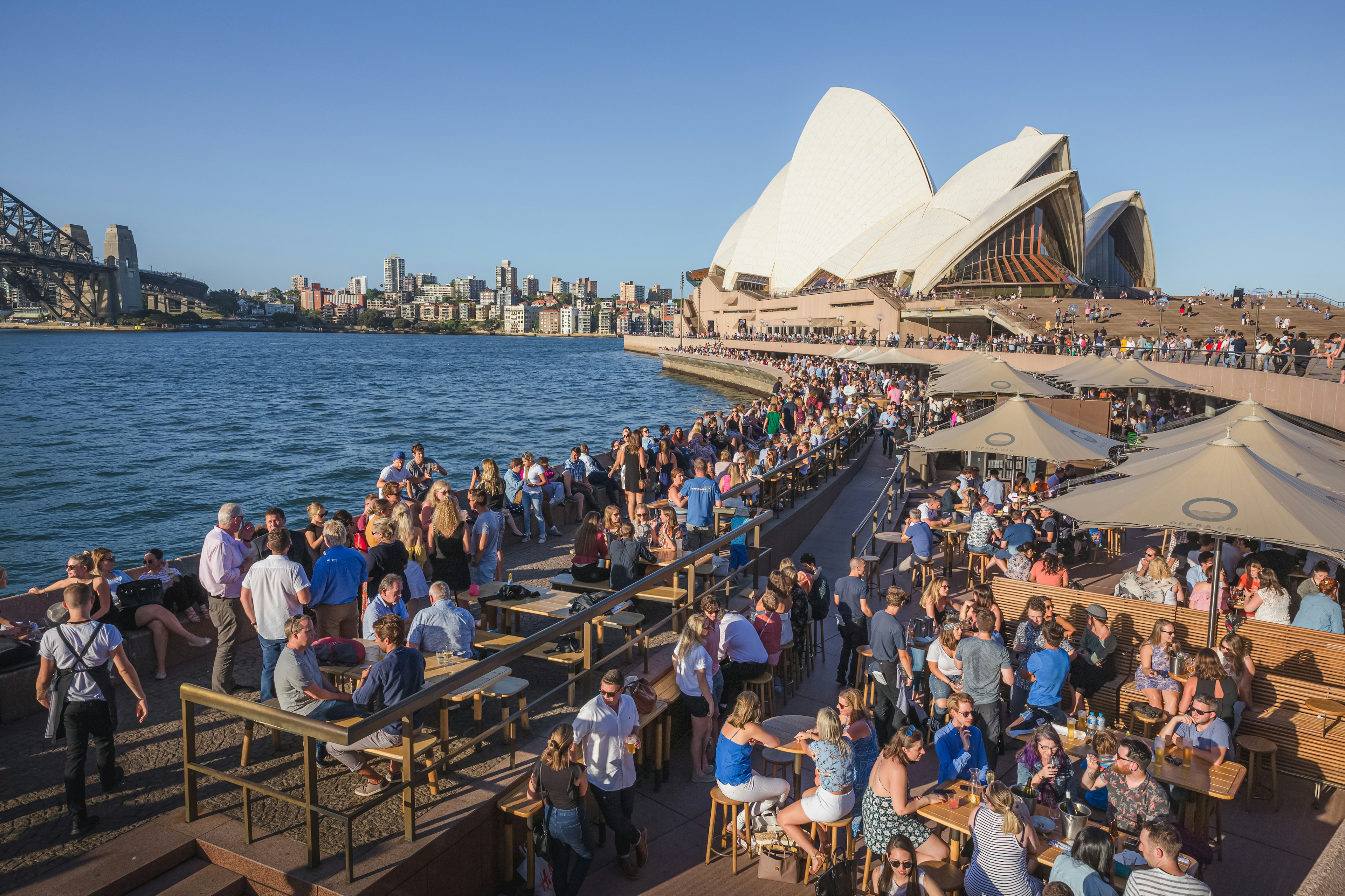 People socialize at picnic tables on a rooftop near Sydney Harbor overlooking the opera house.