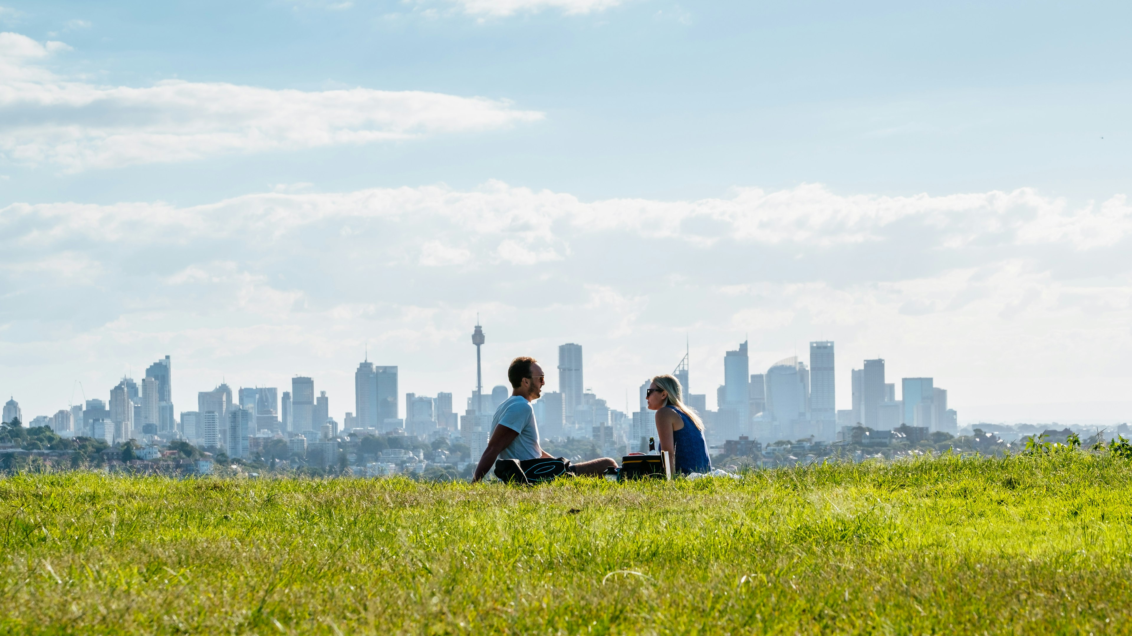 A couple sitting in a field with Sydney's high-rise skyline in the background.