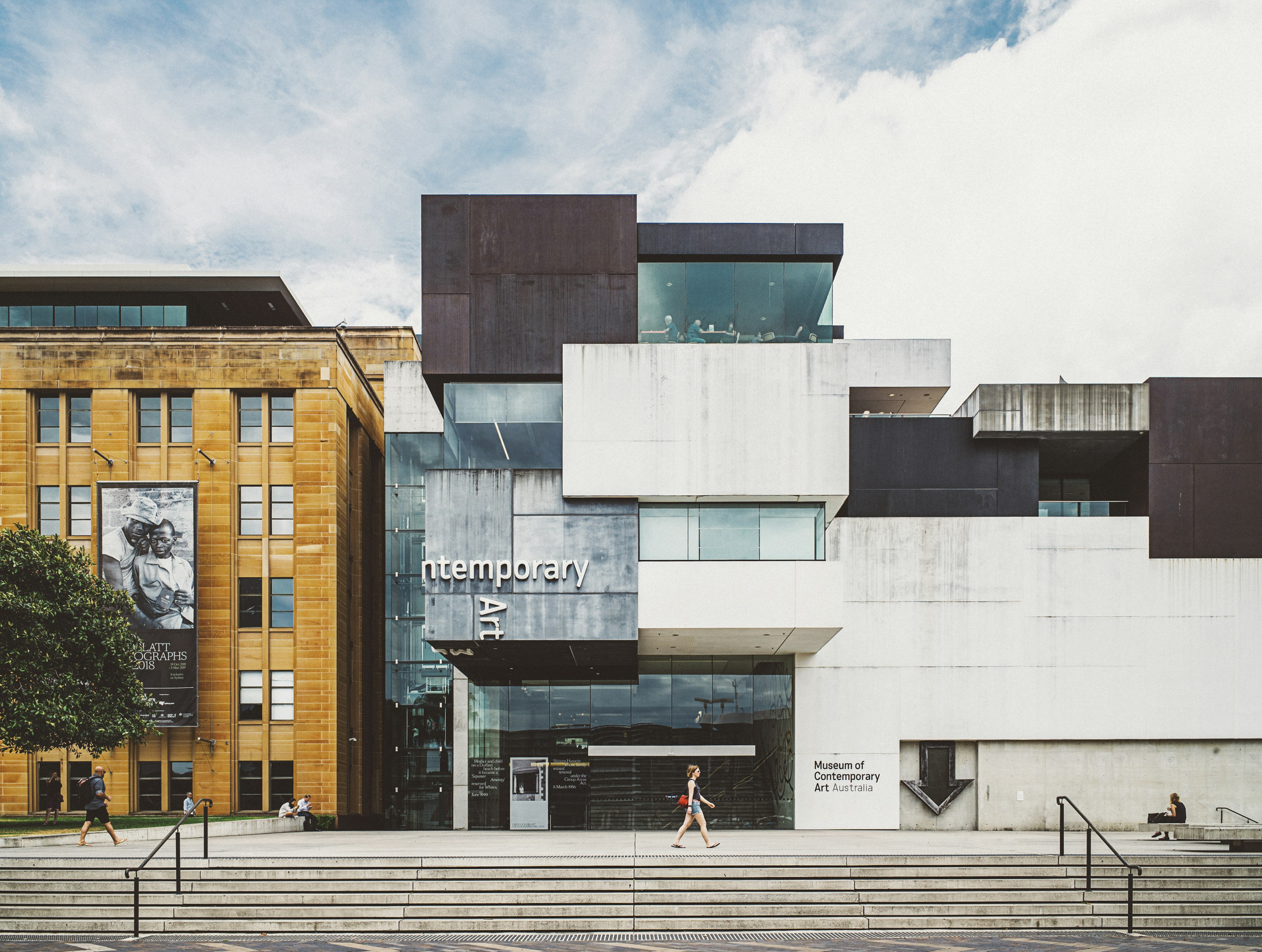 The front steps of the Museum of Contemporary Art Australia (MCA), which is a modern structure with large windows. A woman walks past.