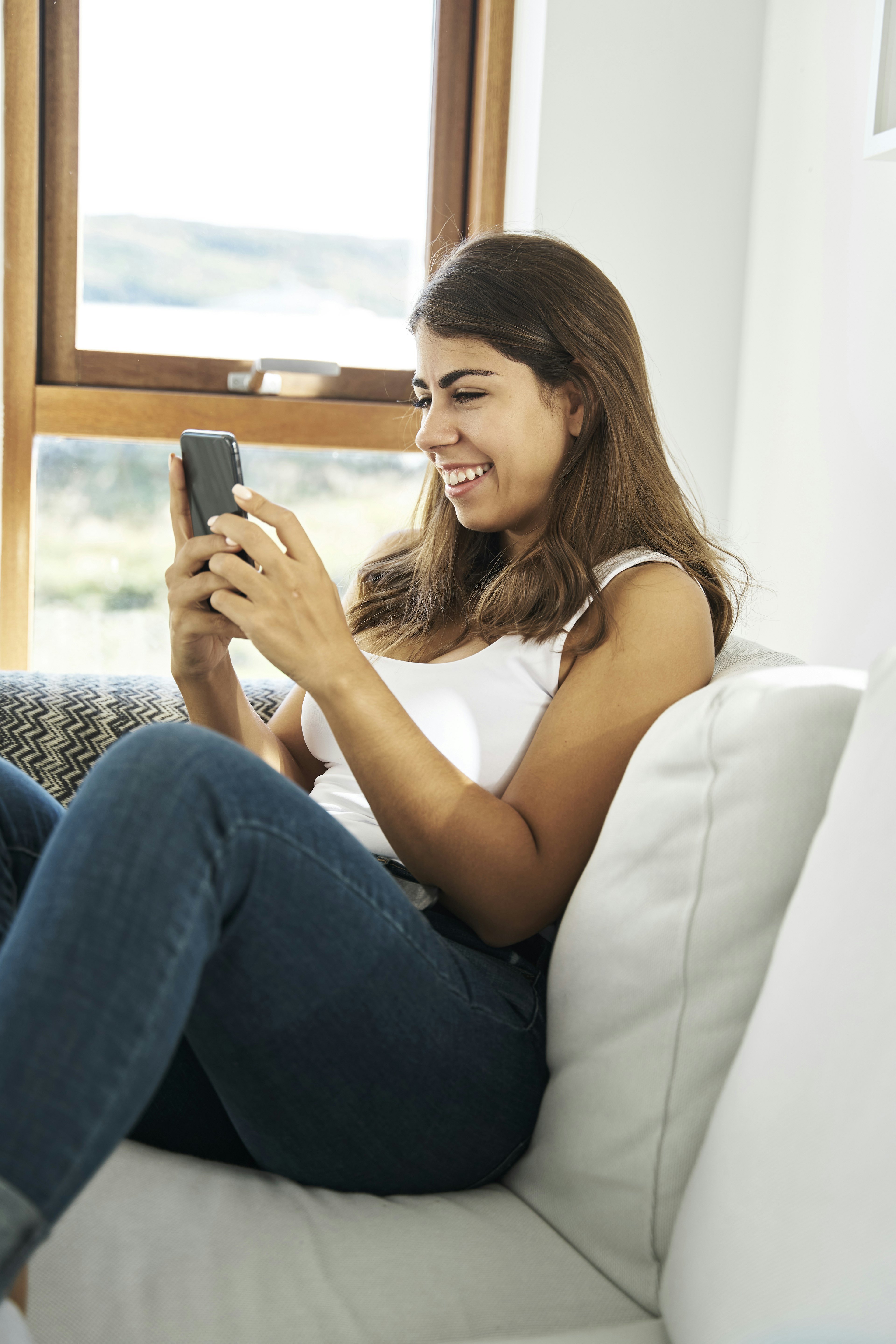 Cheerful young ethnic female in casual clothes sitting on cozy sofa near window and browsing mobile phone while resting at home