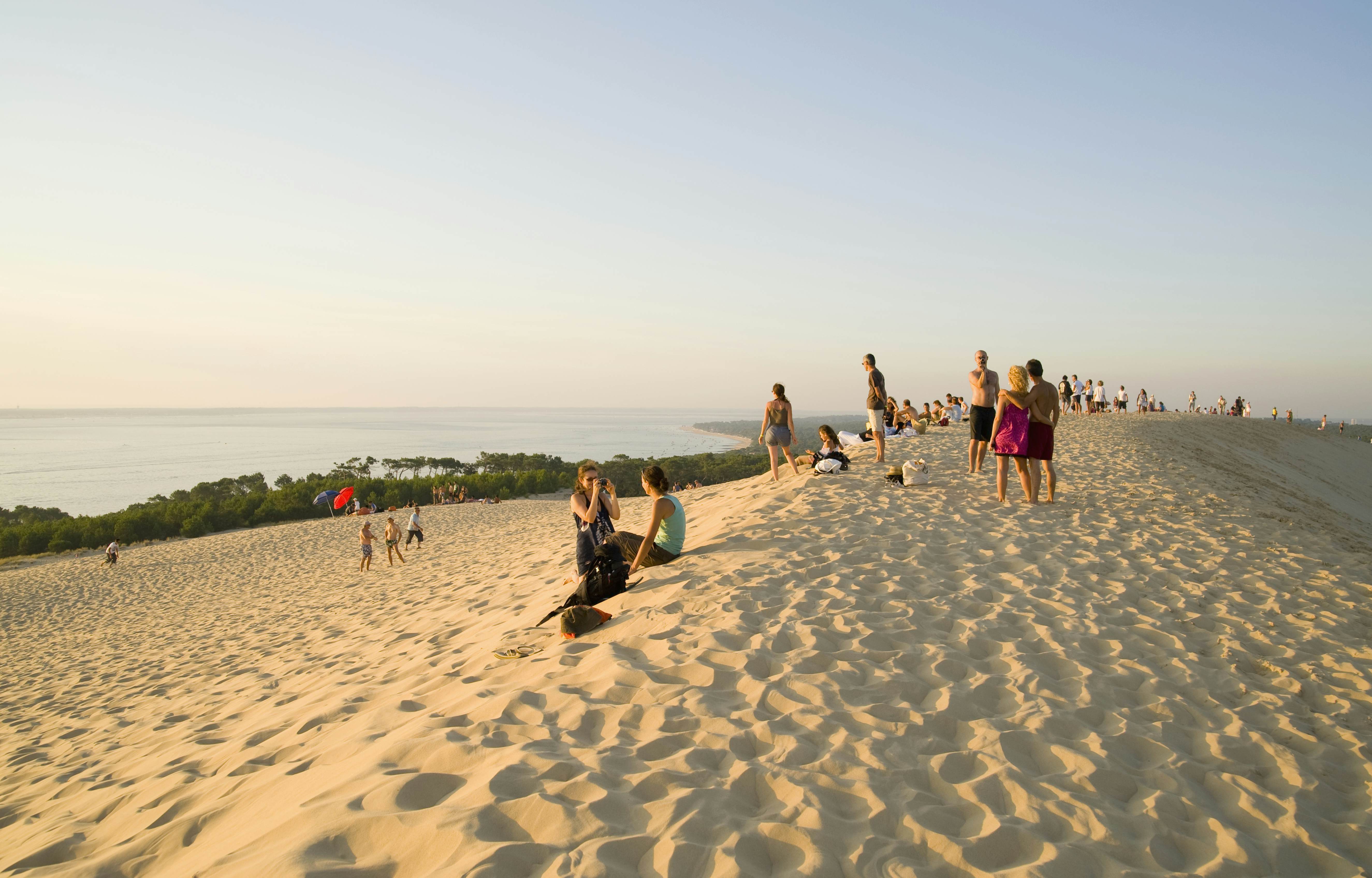 People on the ridge of a giant dune by the sea at sunset.