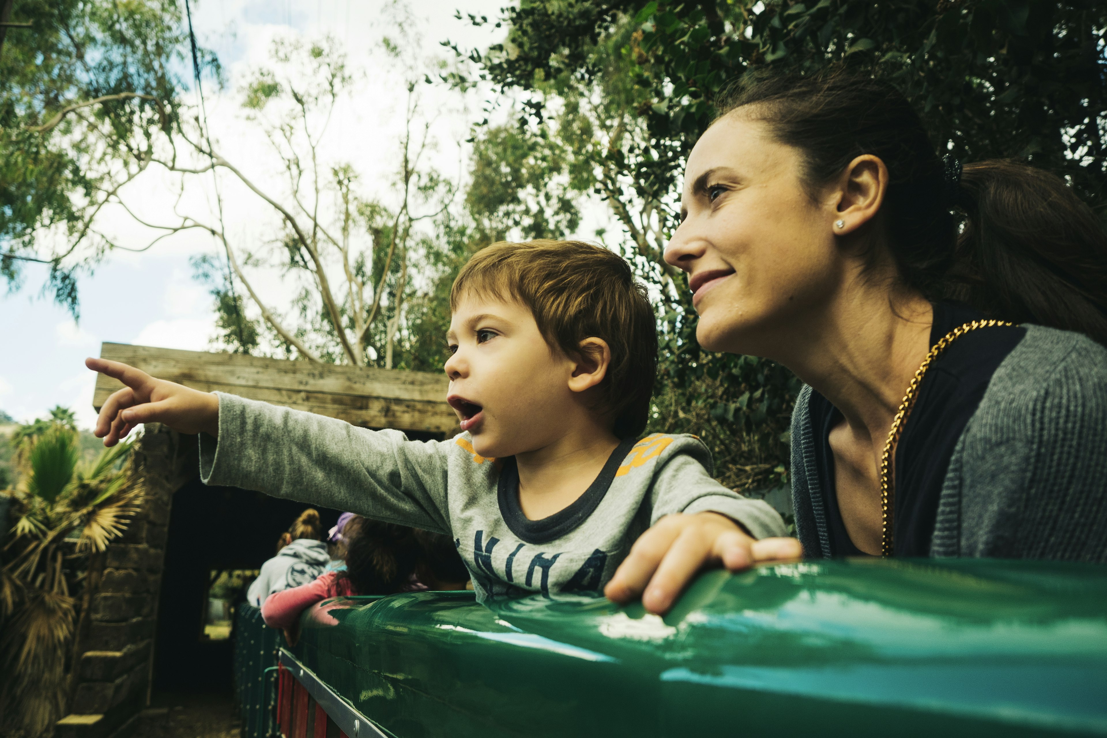 A child riding a train points excitedly at the passing landscape as his mother looks on