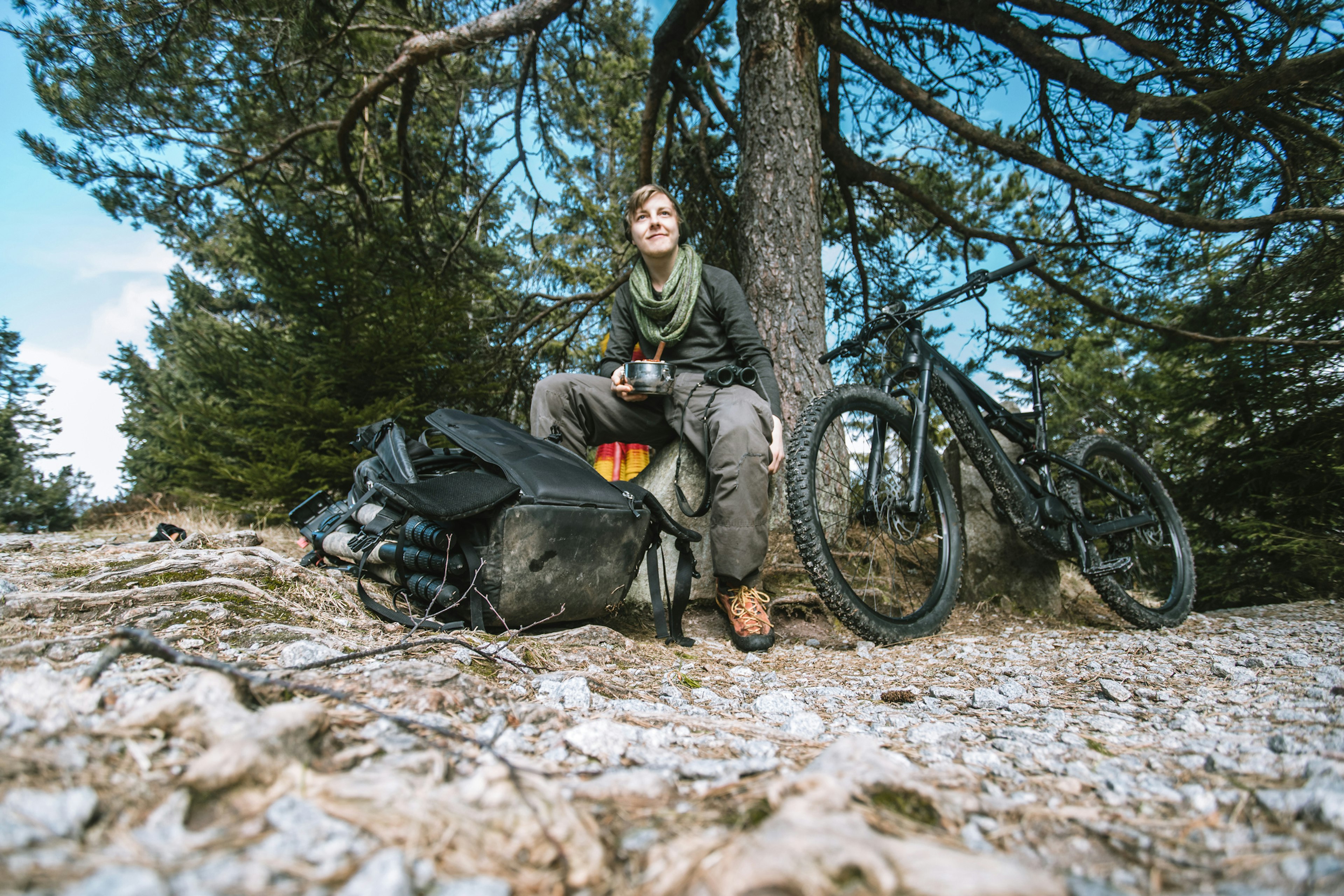 A mountain biker sits on a rock with their gear and bicycle on the forest floor next to them