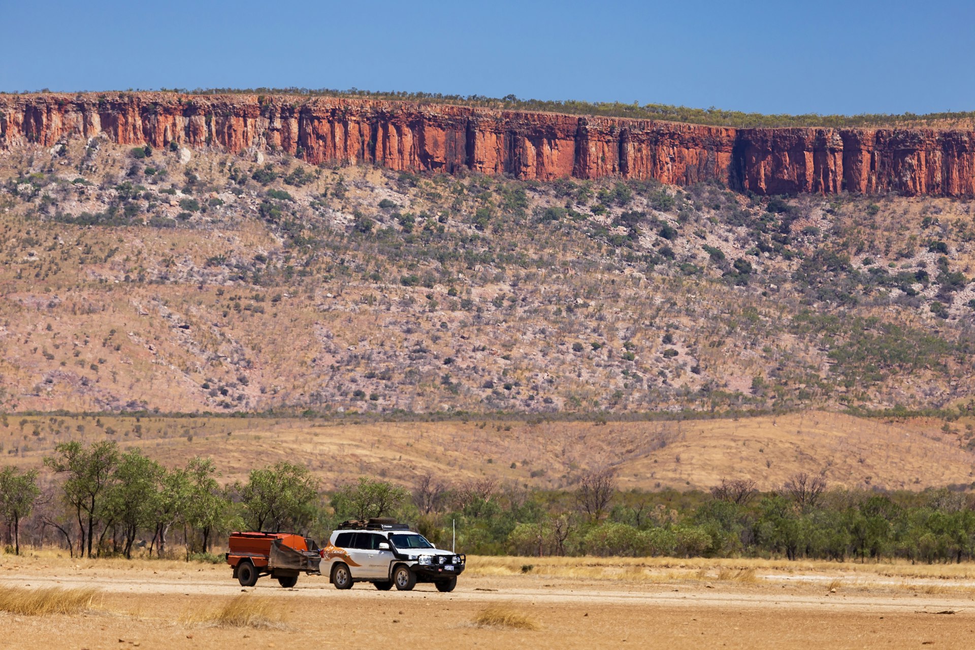 A 4WD vehicle pulling a trailer drives the Gibb River Road with spectacular cliffs in the distance, Western Australia