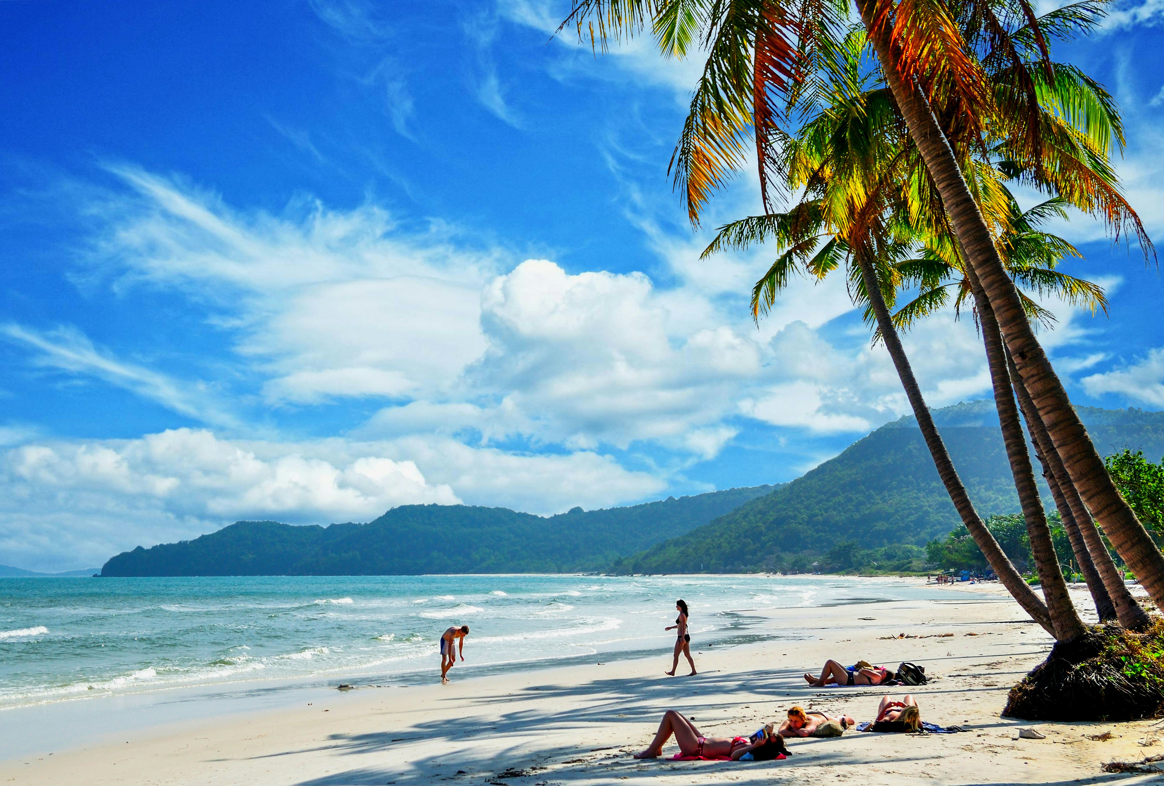 People lie on the sand and walk toward the gentle waves on a beach with white sand and swaying palm trees, with green hills and clouds in the sky in the distance.