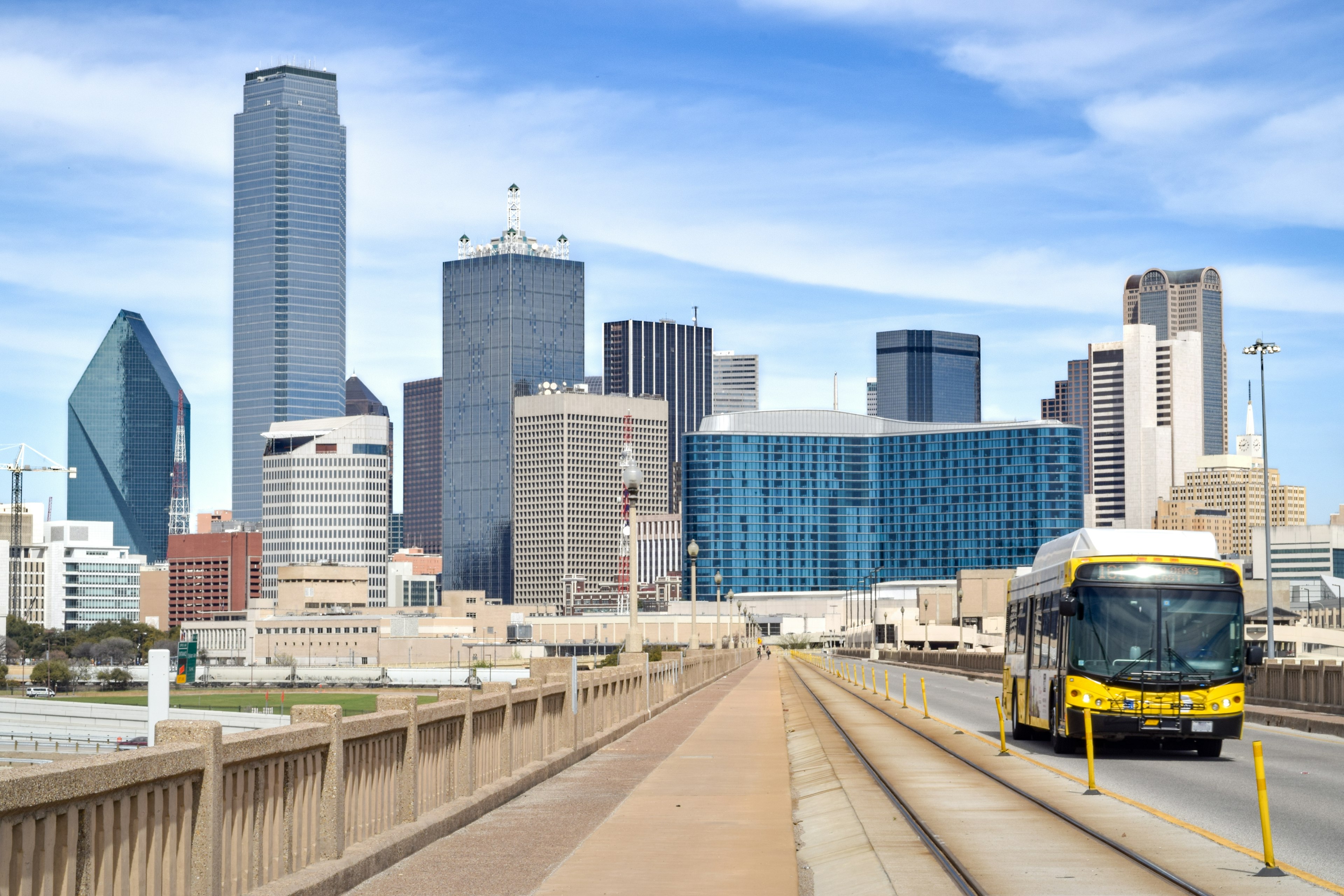 A public bus drives along a highway away from central Dallas, Texas. In the background a number of skyscrapers are visible.