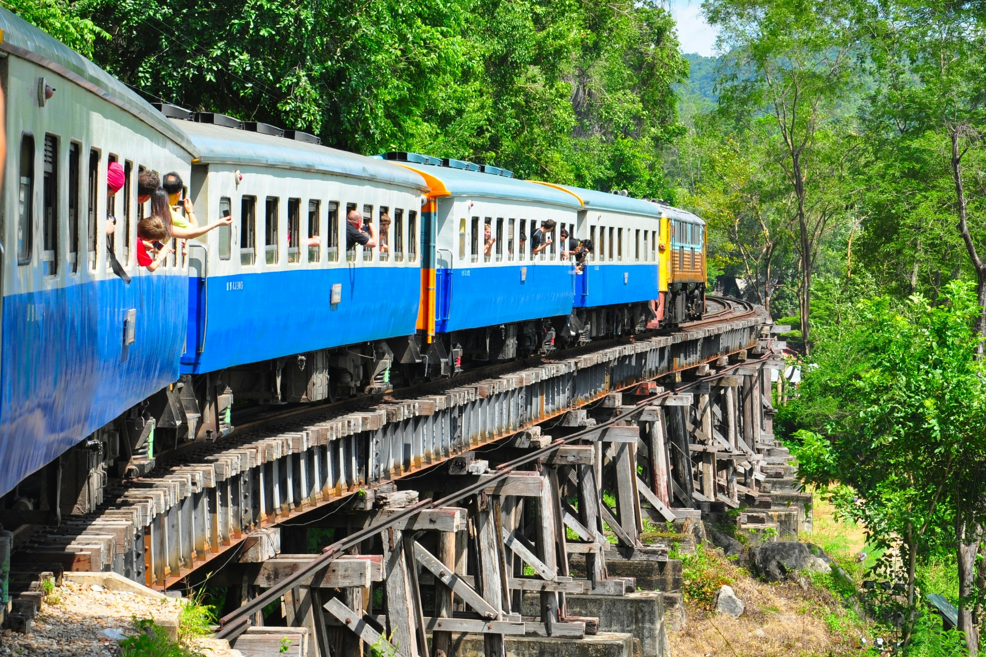 Tourists lean out of a train window as it travels over a bridge through the Thai countryside