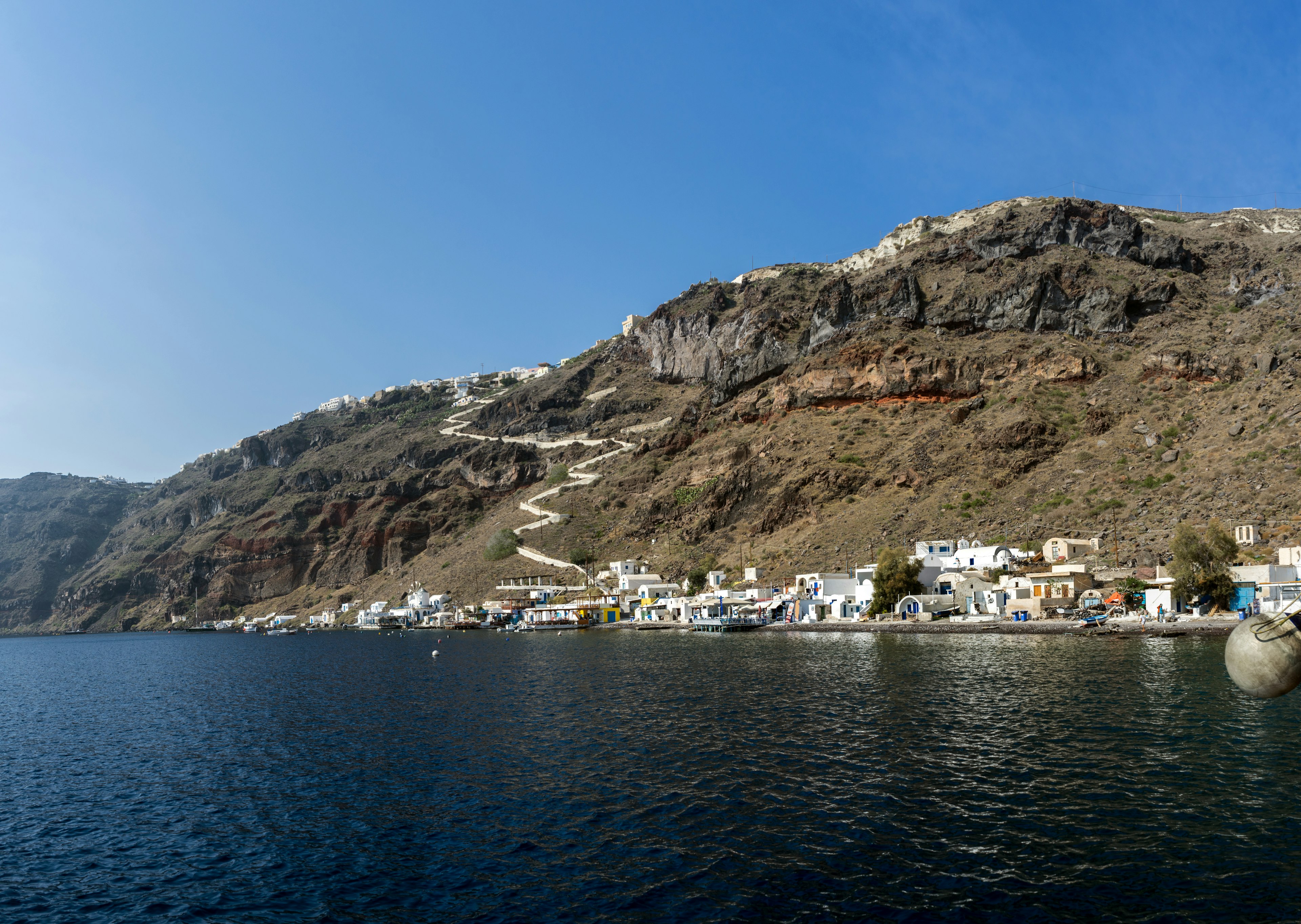 A shot taken from sea towards a volcanic island, with whitewashed houses lining the shore. A winding path weaves down the hill towards the village