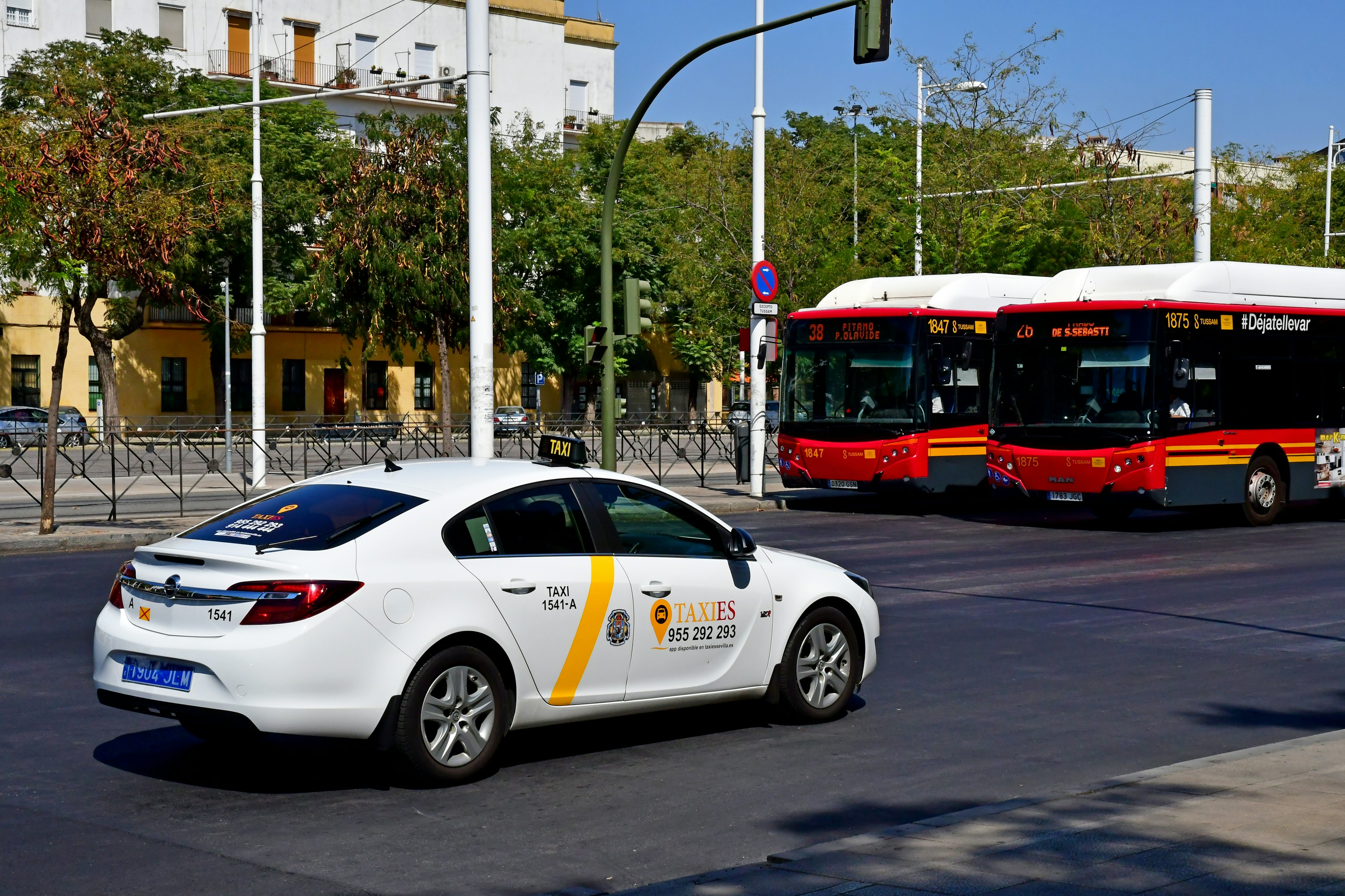 A white taxi with a diaganol yellow stripe, and two red buses waiting at a signal