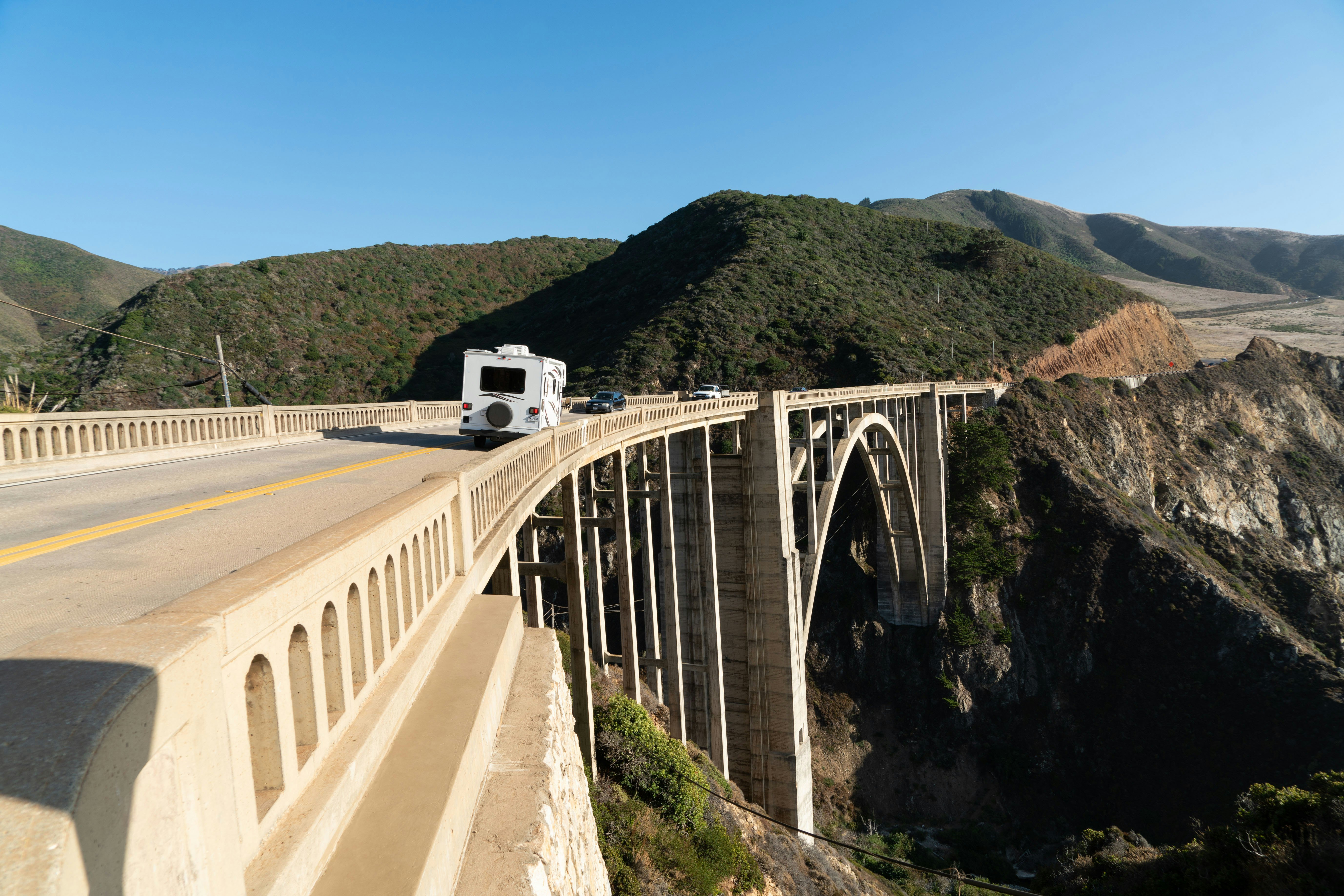 A camper van crosses a bridge with a large arch that spans a deep creek.