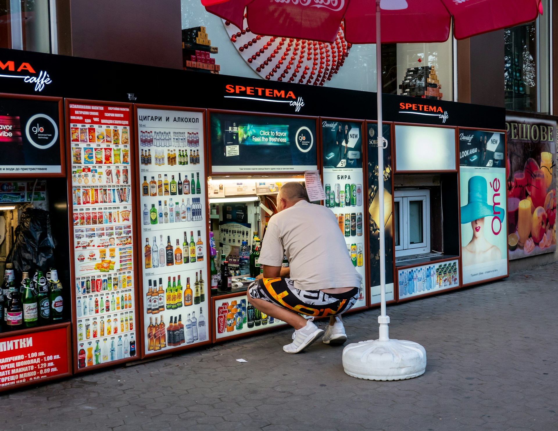  man in shorts and t-shirt squats down to a very low level shop where the assistant serves alcohol and tobacco from a basement window, Sofia, Bulgaria