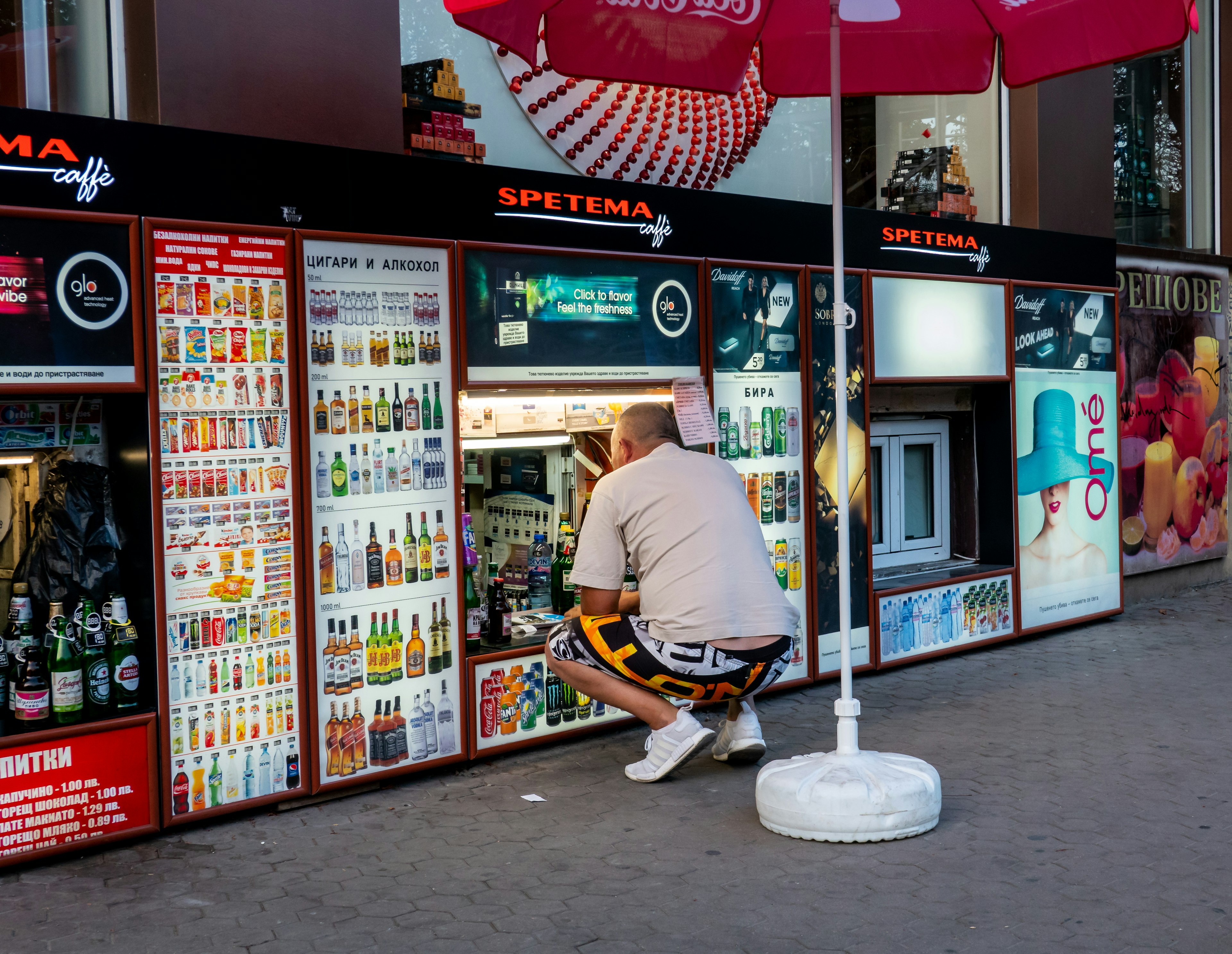 man in shorts and t-shirt squats down to a very low level shop where the assistant serves alcohol and tobacco from a basement window, Sofia, Bulgaria