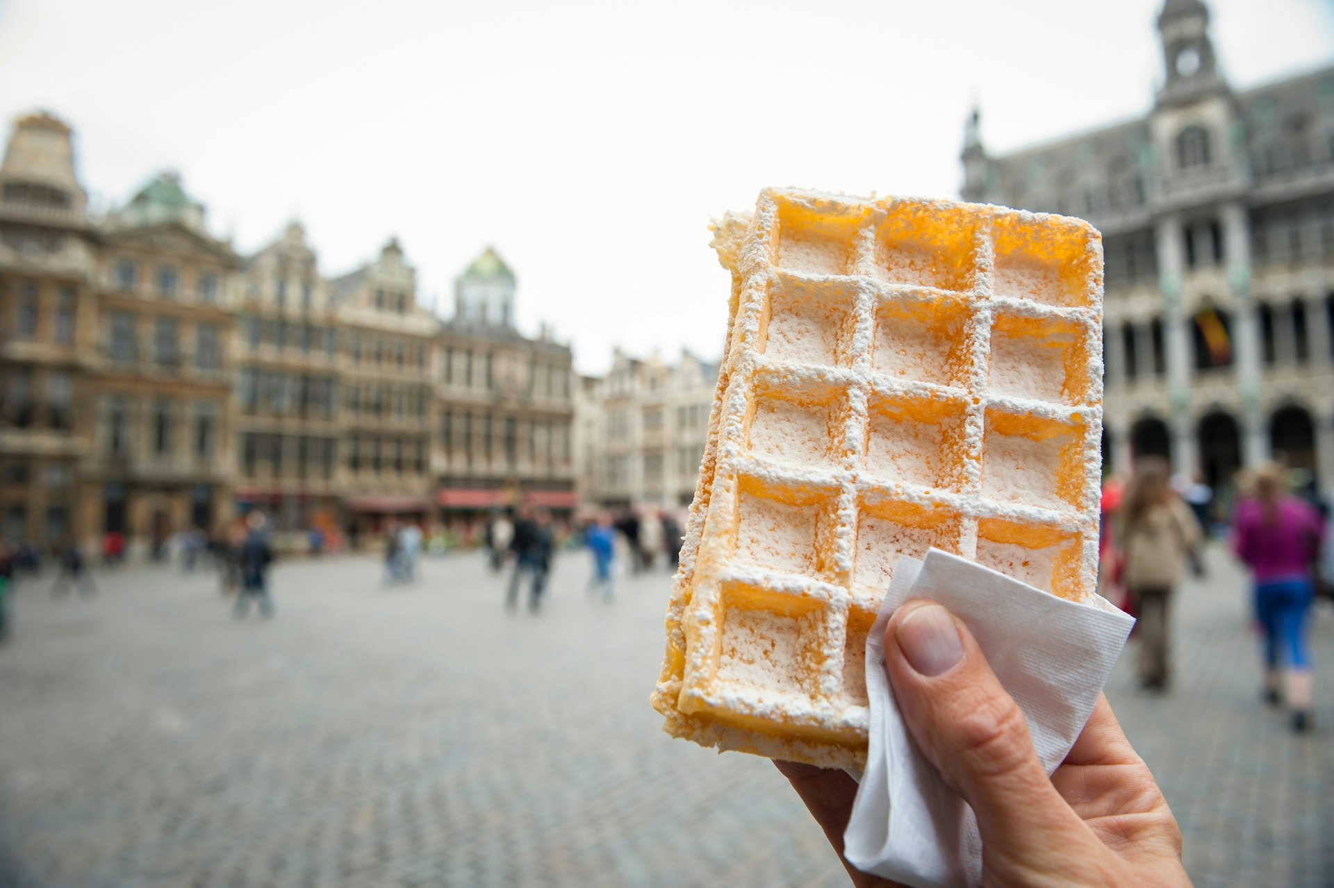 Hand holding Belgian waffle dusted with powdered sugar outdoors in the Grand Place in Brussels