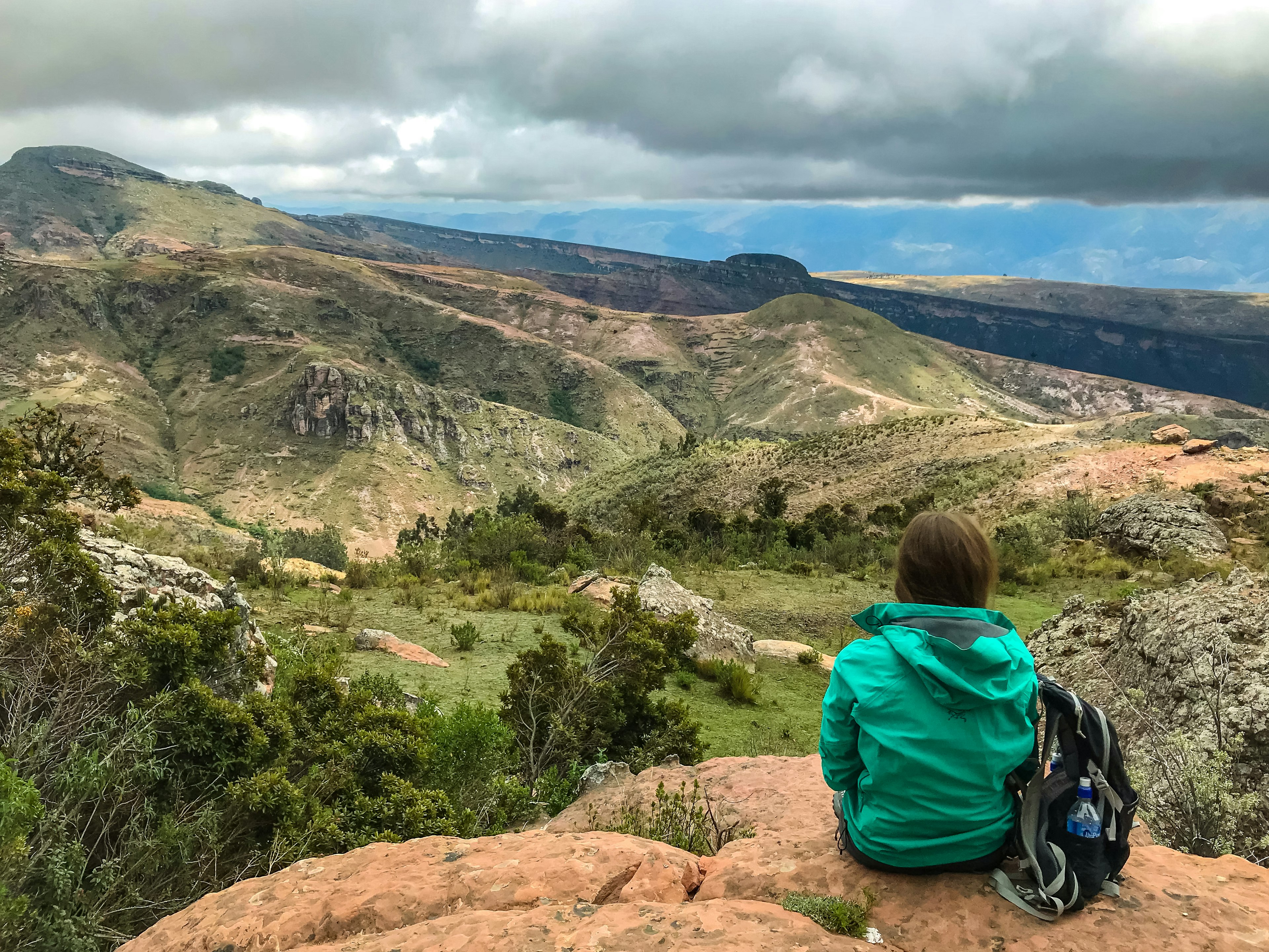 Woman with a green raincoat shot from behind, sitting at the edge of a rock point looking at the landscape at Torotoro National Park, Potosí, Bolivia