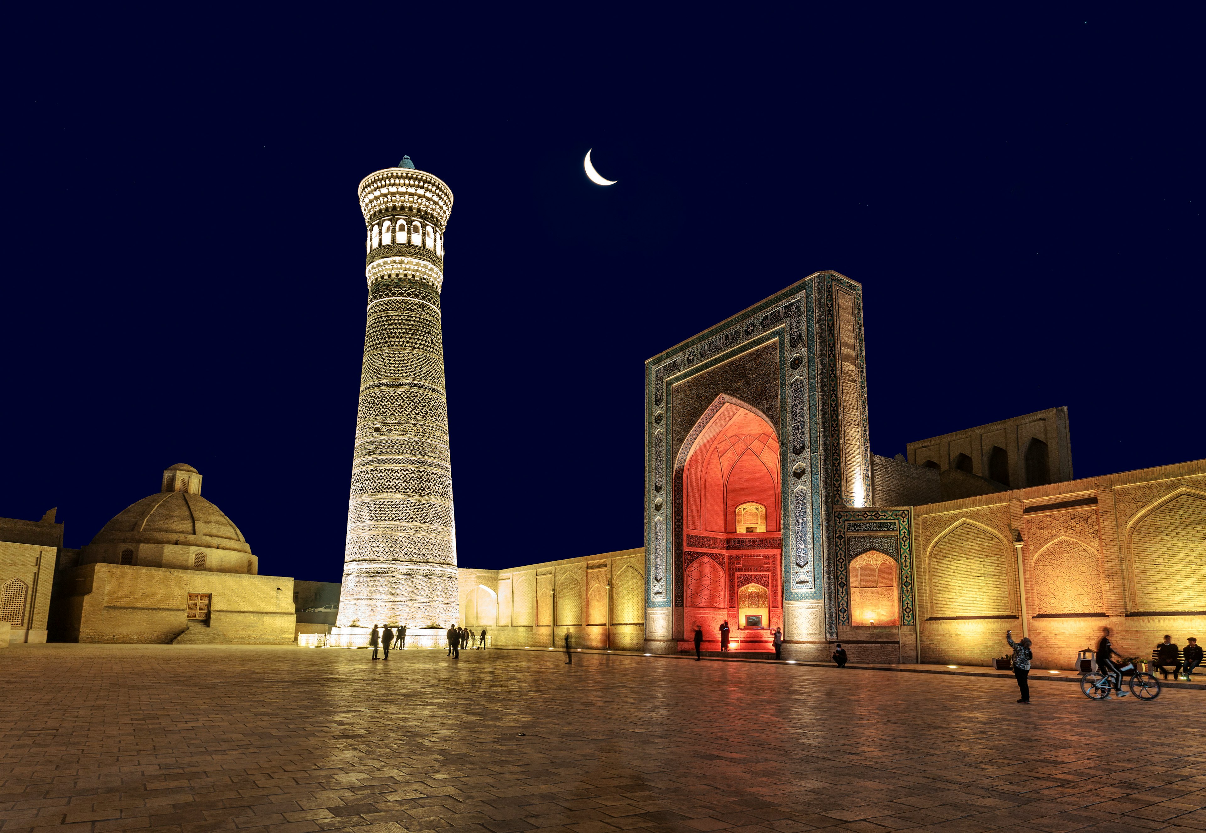 An Uzbek minaret and mosque in Bukhara, Uzbekistan are lit up at night with a crescent moon above.