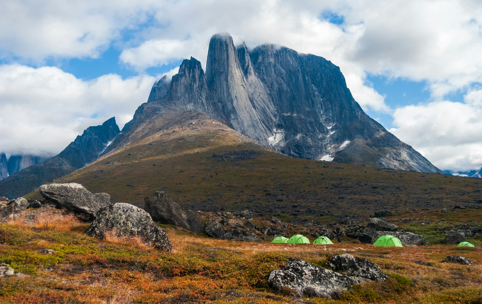 Lime green tents sit in a field at the base of a sheer, gray mountain in the Tasermiut Fjord