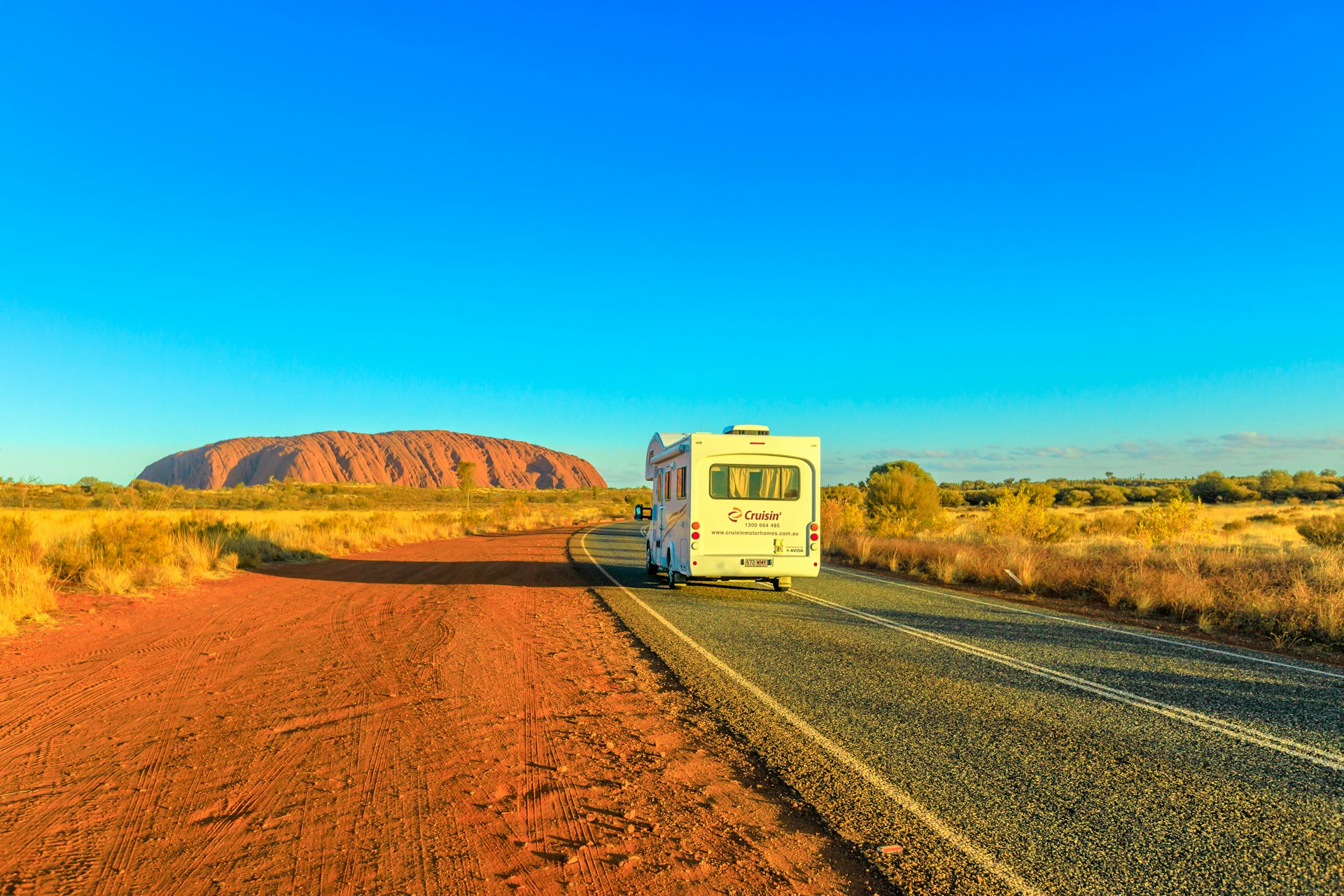 A camper van drives on the road leading to Uluru (Ayers Rock) at sunset, Northern Territory, Australia