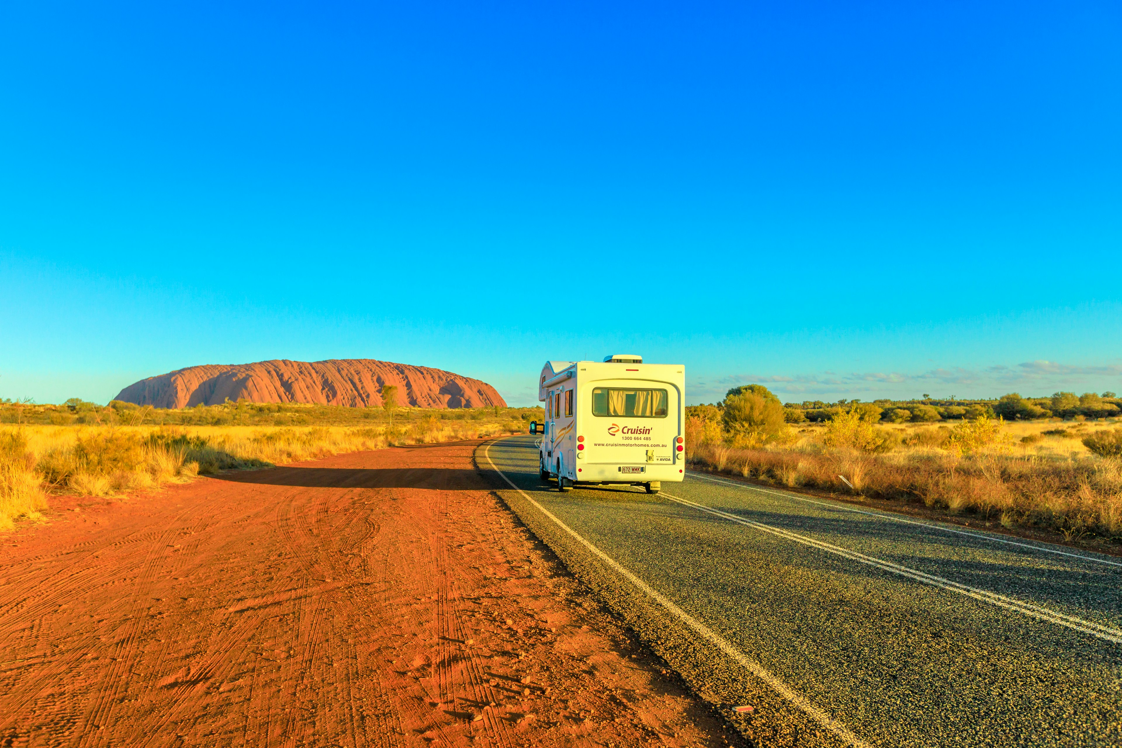 A camper van drives on the road leading to Uluru (Ayers Rock) at sunset, Northern Territory, Australia