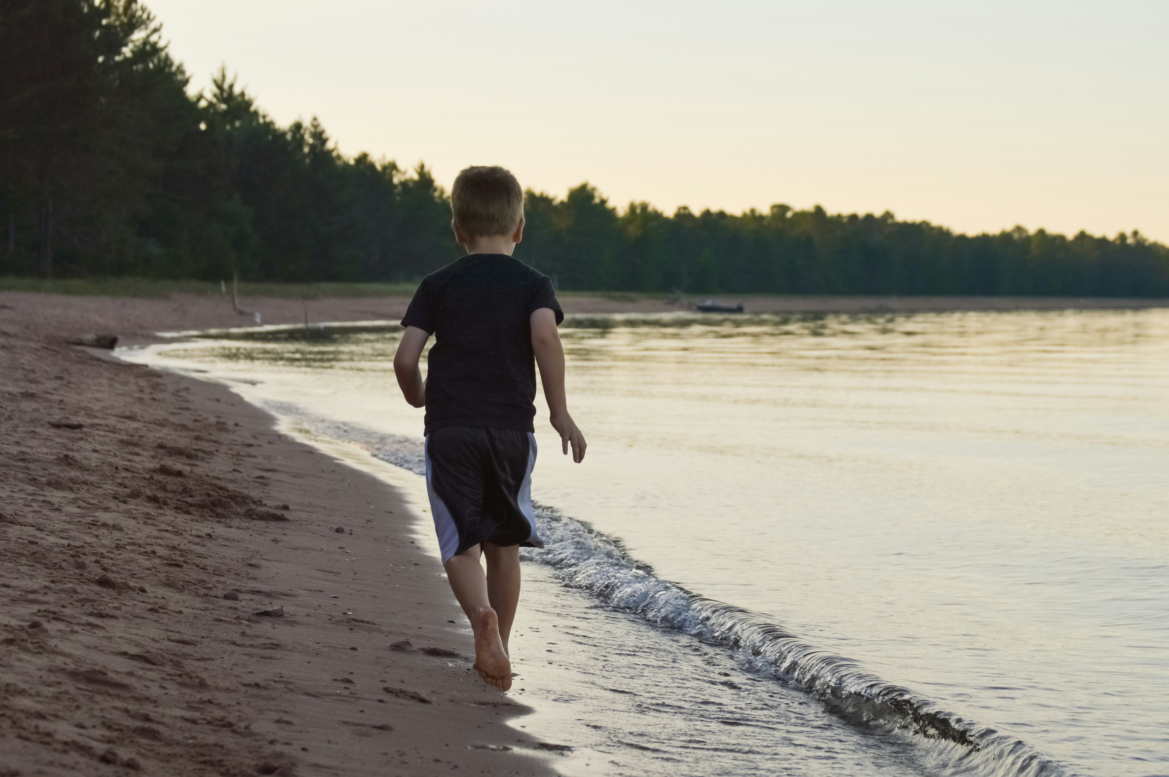 School aged boy runs on a deserted sandy beach surrounded by evergreen forest with a boat beached in the distance under yellow evening sky, Northern Wisconsin, Great Lakes, USA.