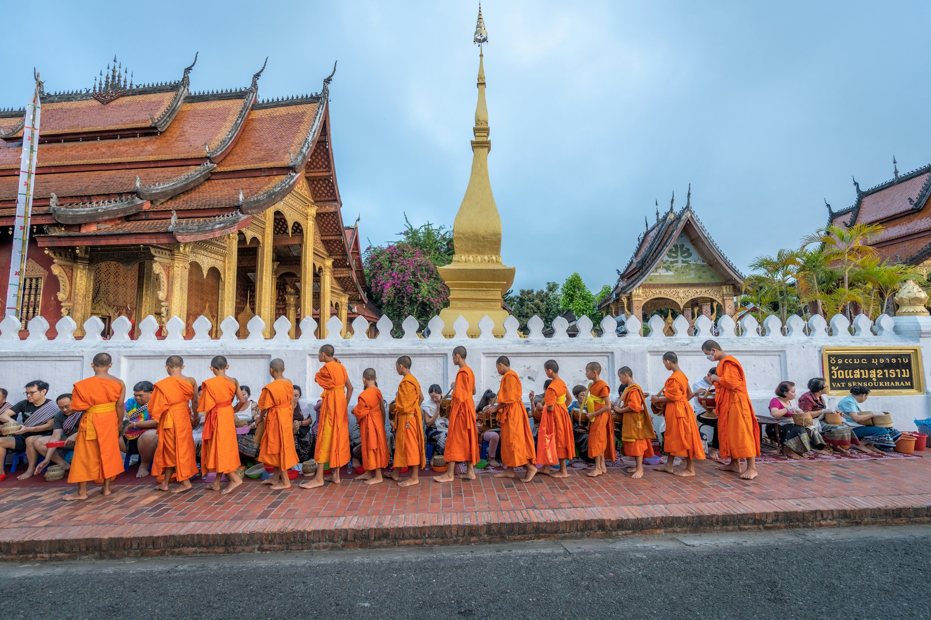 Monks in orange robes line up to take alms in front of a golden stupa on a street in Luang Prabang, Laos