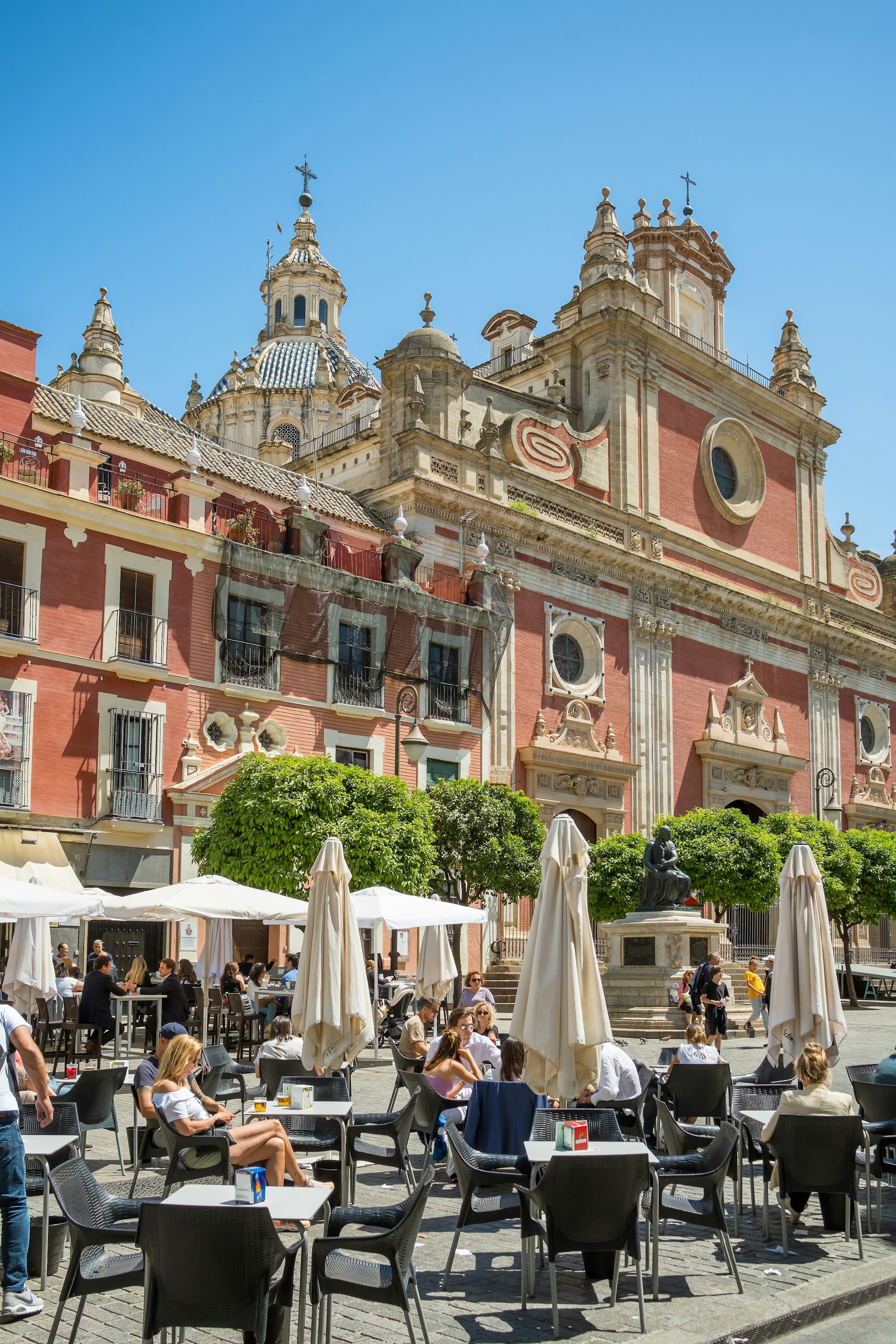 A series of tables with sunshades in front of a large baroque church building, with a pale-pink facade and many turrets