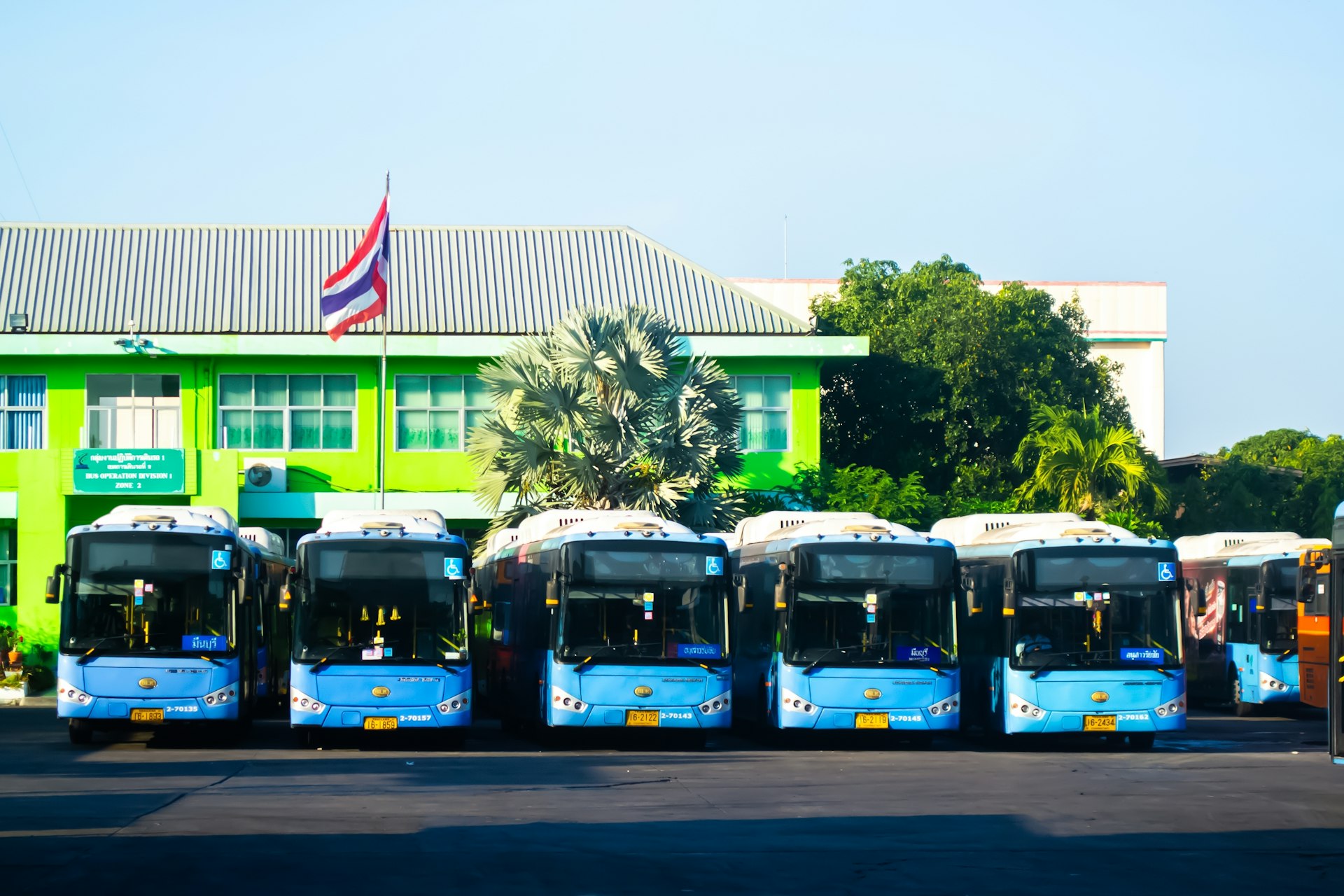 A row of blue buses lined up outside a building with the red, white and blue striped Thai flag flying above