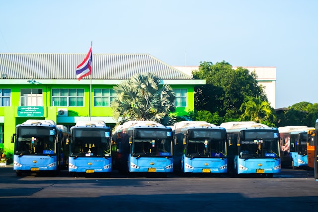 A row of blue buses lined up outside a building with the red, white and blue striped Thai flag flying above