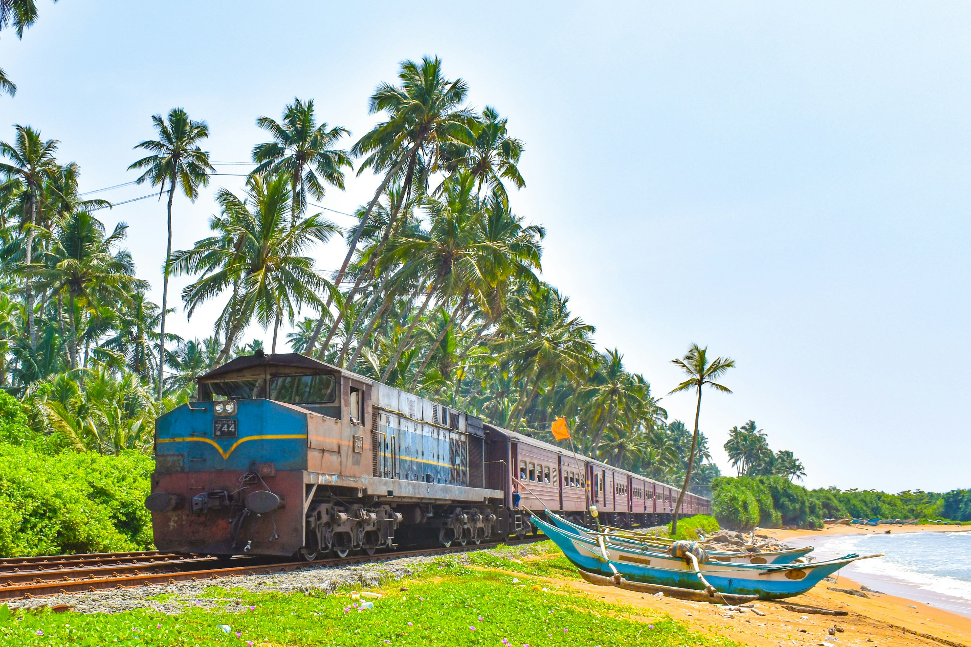 A diesel passenger train passes a beach with blue fishing boats on a coastal track lined with palm trees