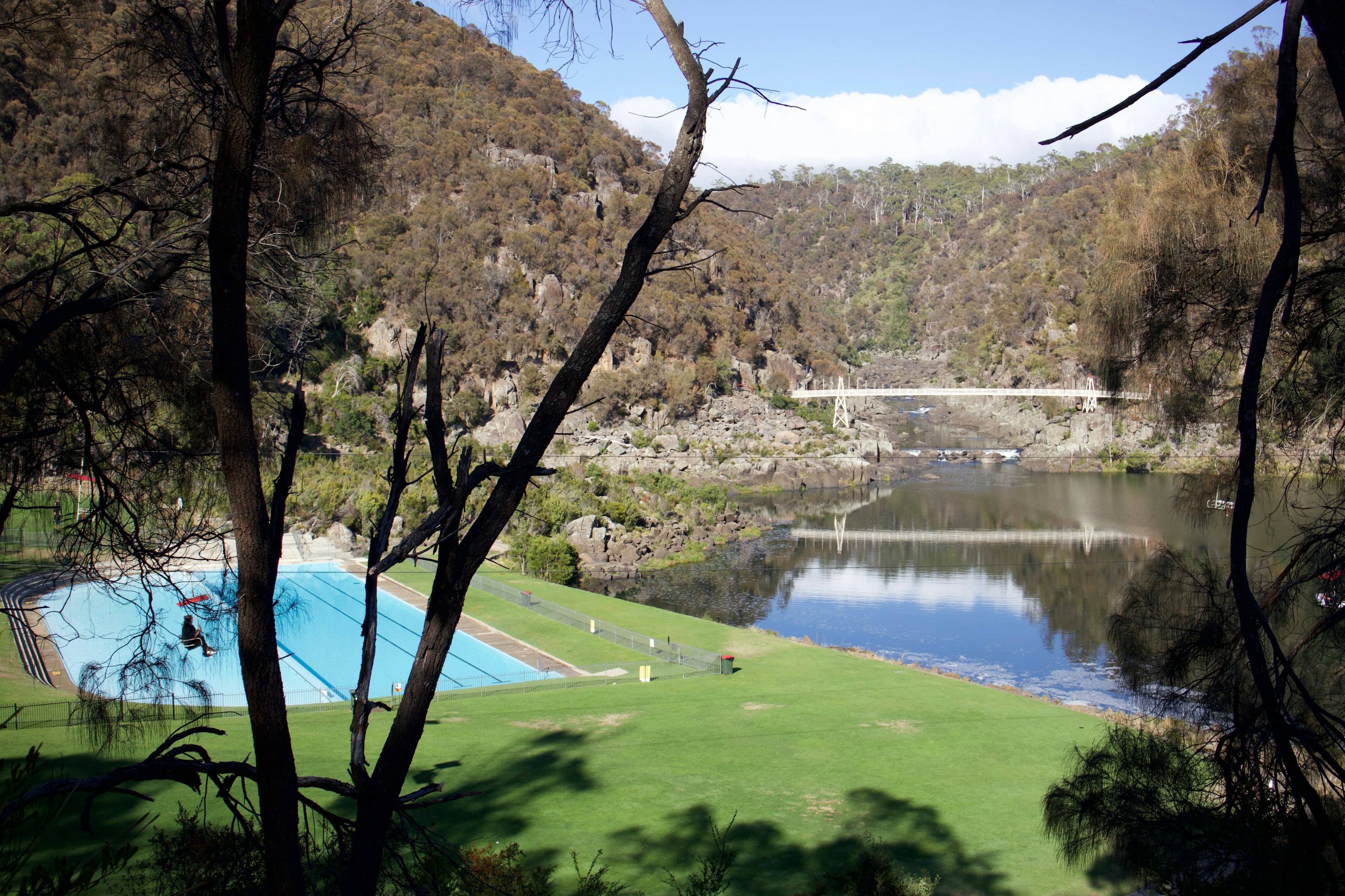 A scenic view of a grassy area featuring a swimming pool, surrounded by trees and a tranquil river. A bridge and hills are visible in the background, creating a serene landscape