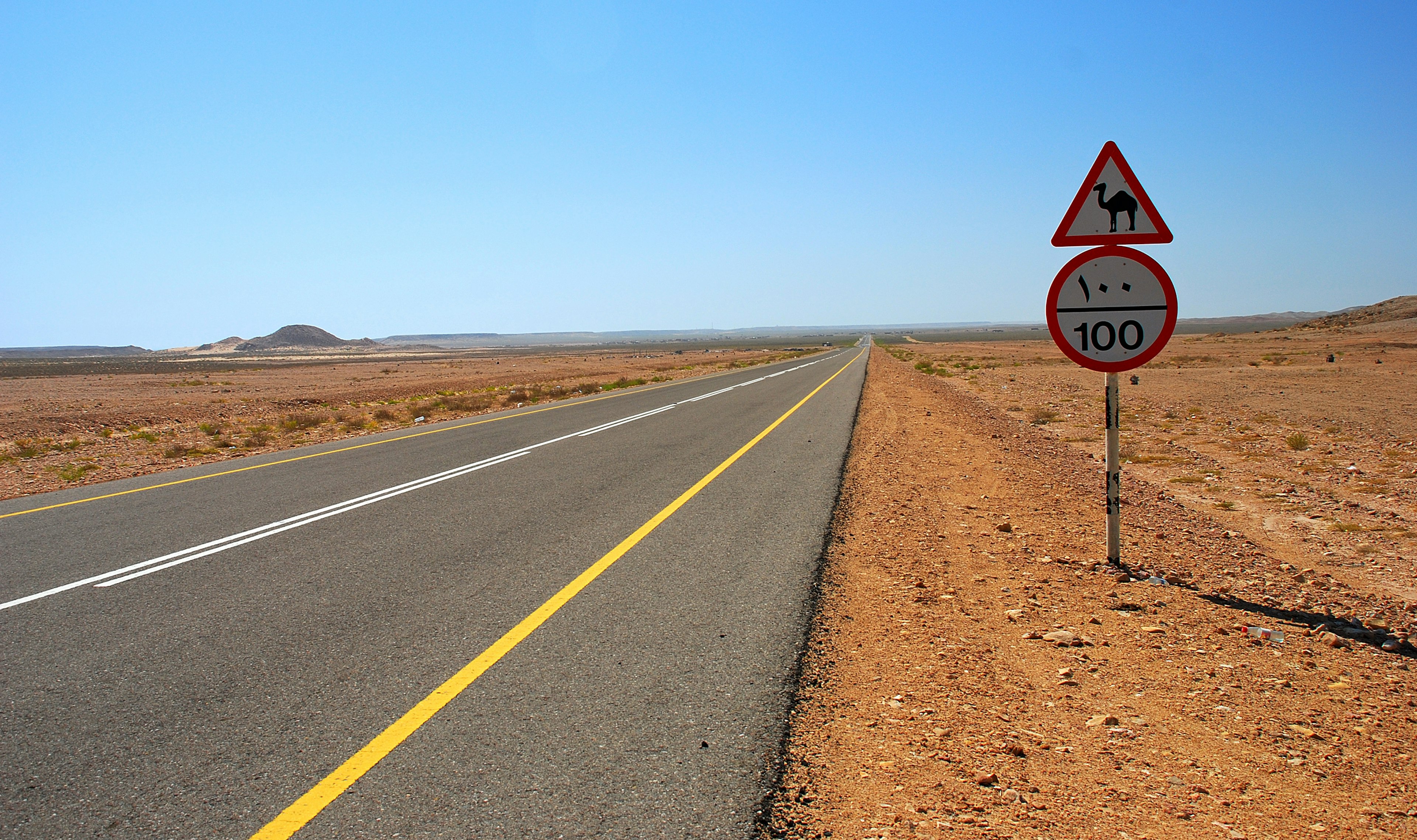 An empty paved road (with a camel crossing caution sign by the side) stretches out into the desert of Oman