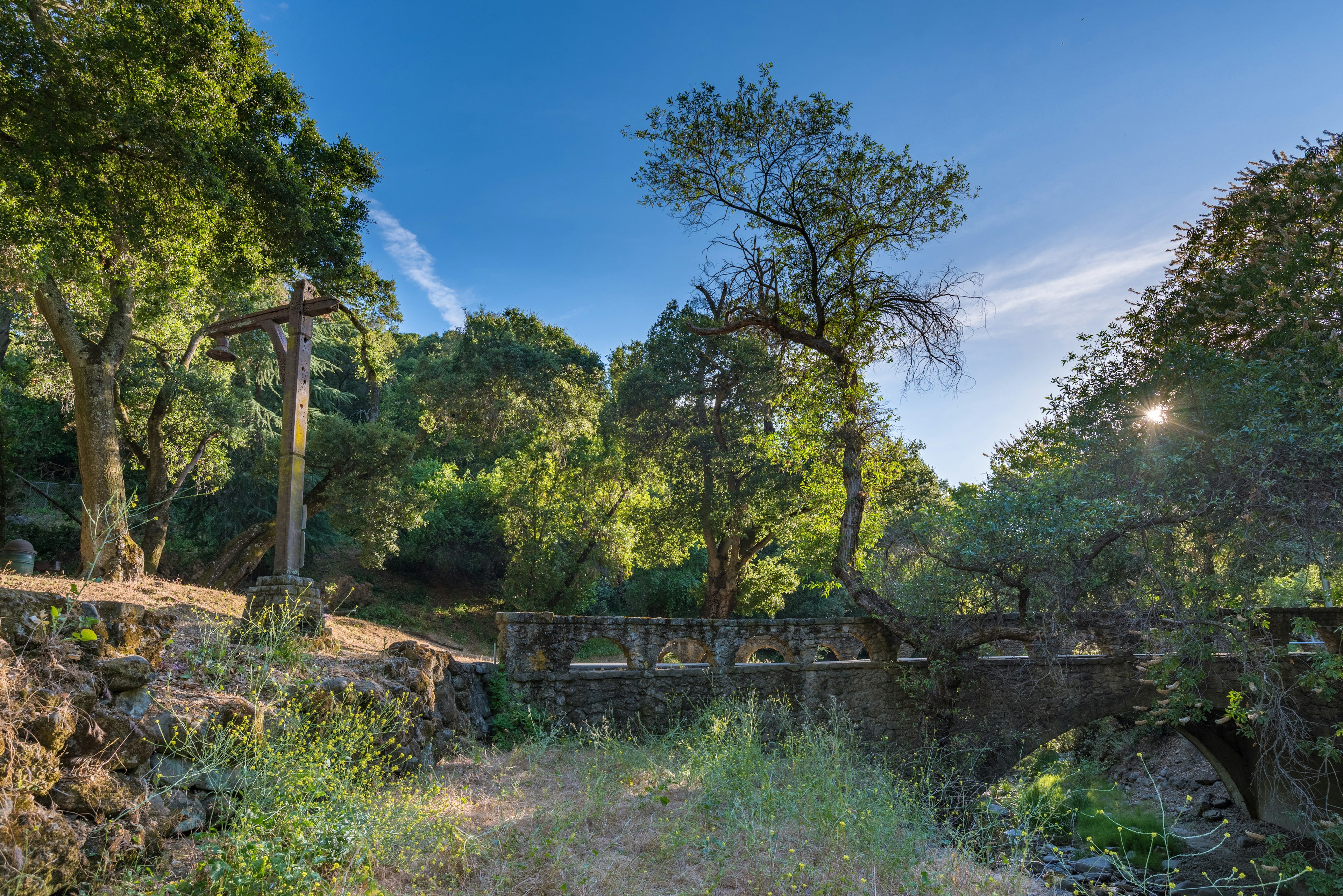 A stone bridge over a gully in a densely wooded area with the sun shining through the trees