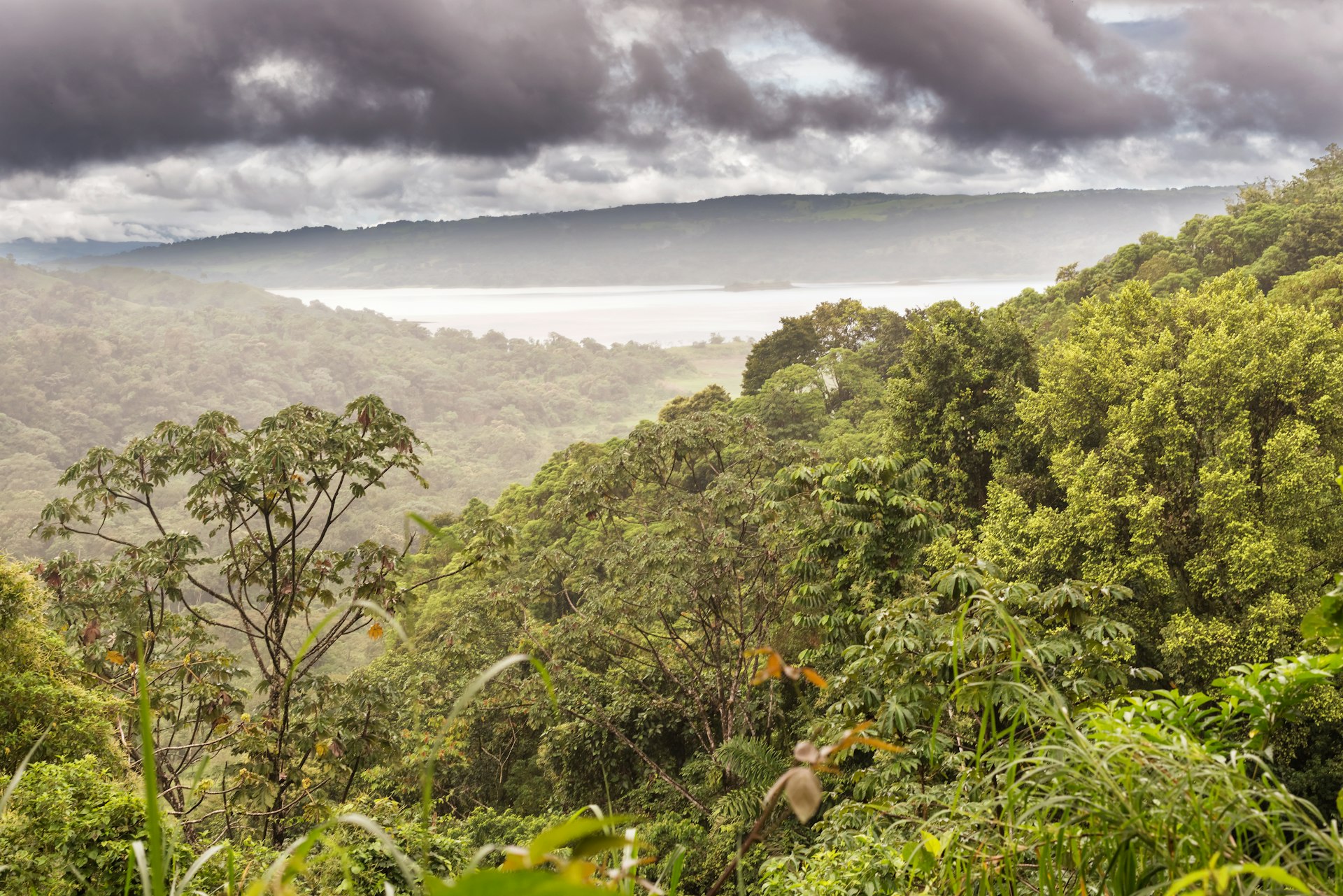 A hillside covered in trees shrouded in mist under dramatic clouds with a view of Laguna de Arenal as seeon from Rancho Margot in Costa Rica