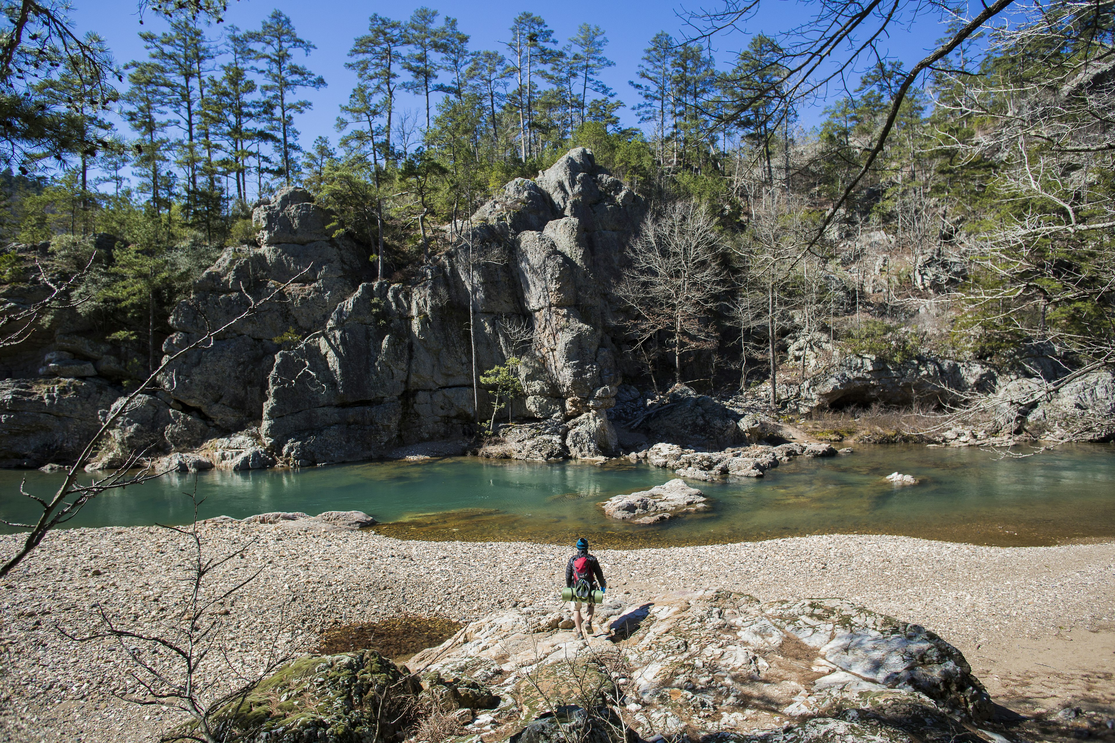 Hiker with full backpacking gear walks towards large rock formations and a river in the Ouachita Mountains in West Central Arkansas