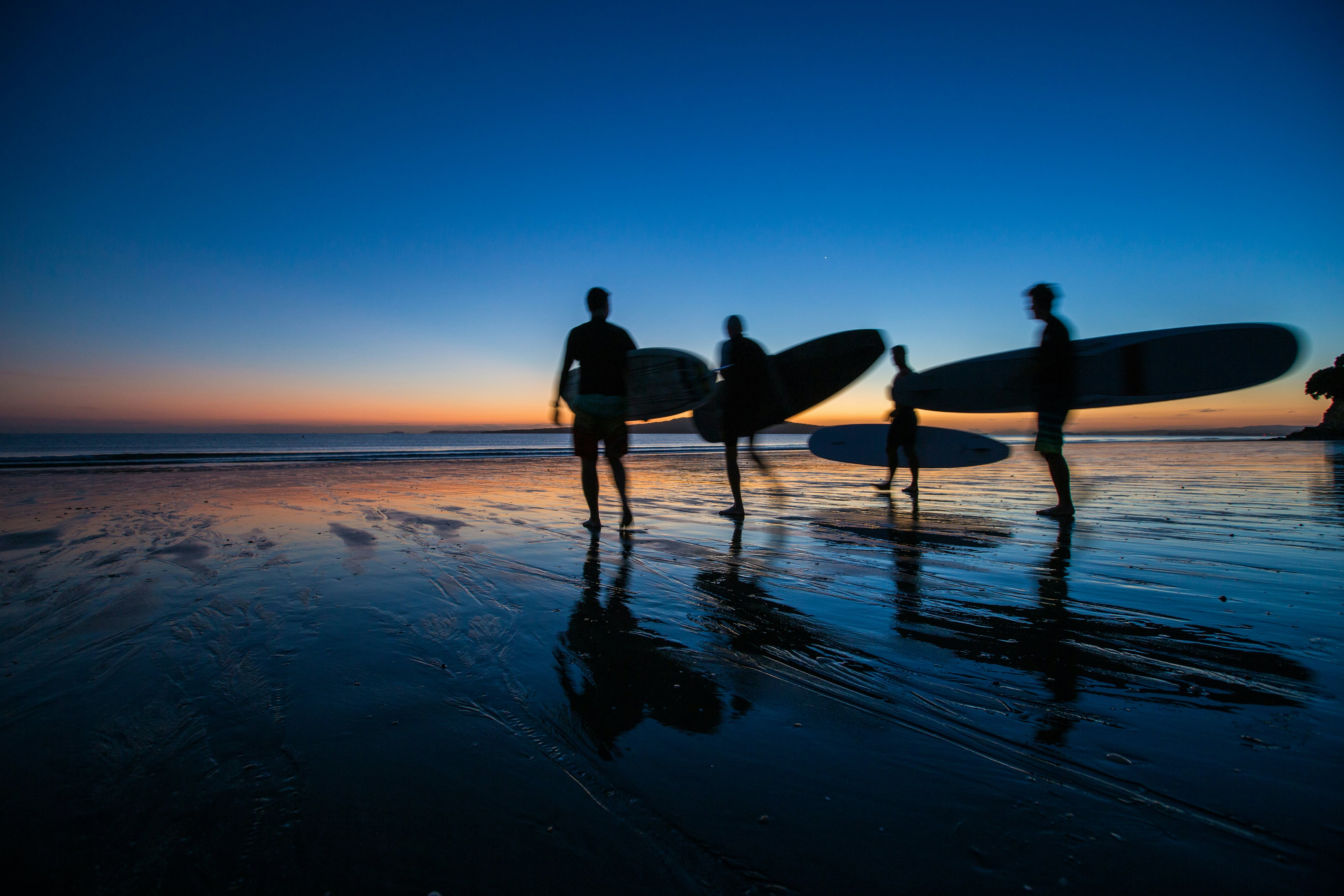 Surfers silhouetted on the beach at dawn with a pink-and-blue sunrise at Takapuna Beach