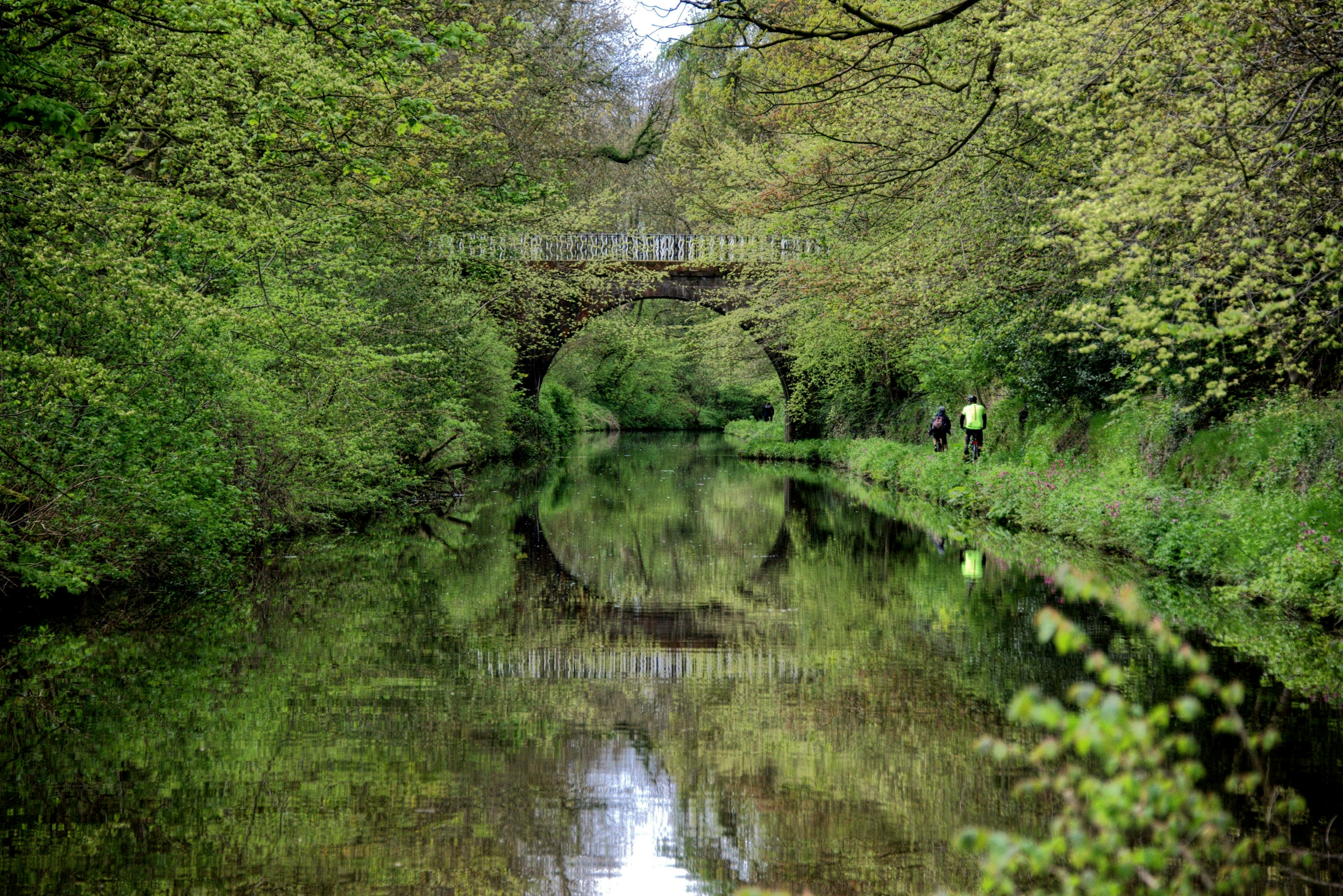 Two cyclists ride bikes along a canal towpath surrounded by greenery. A bridge crossing the canal is reflected in the water