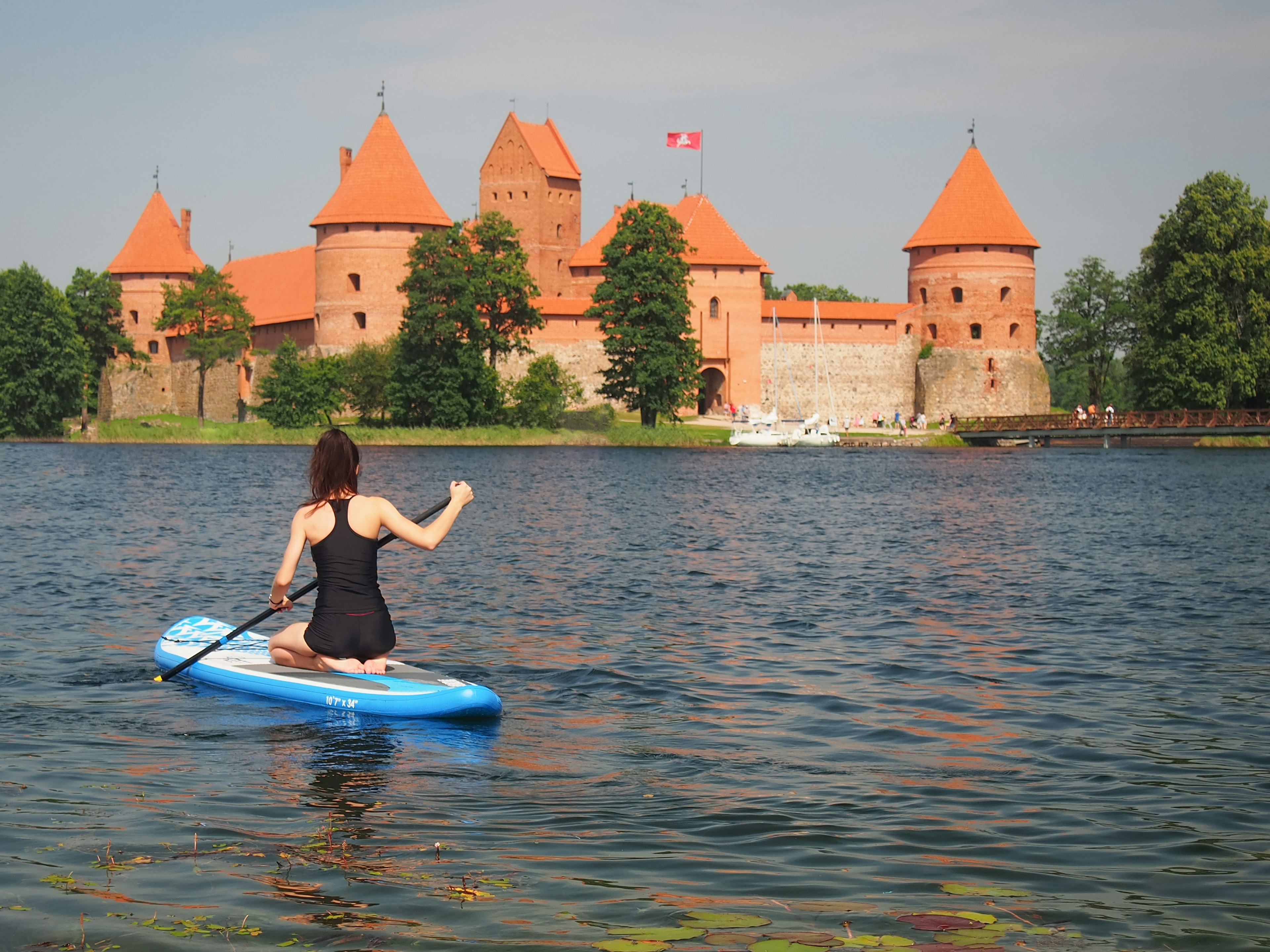 A woman in a bathing suit on a blue stand-up paddle board on Lake Galvė, with Trakai Castle pictured in the distance