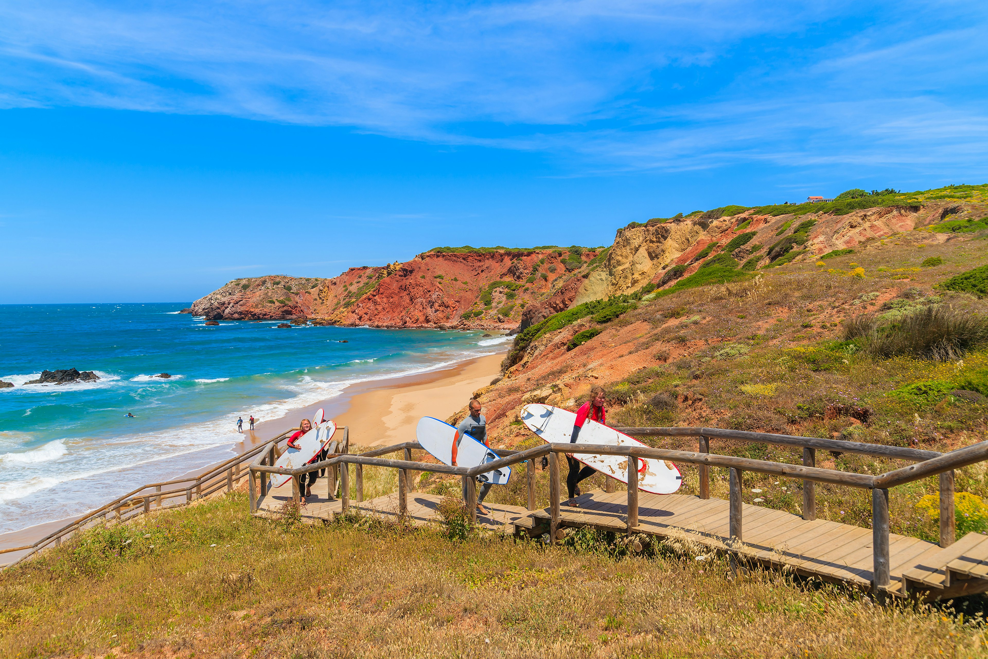 Surfers holding surfboards walk on a footbridge from a rocky, cliff-lined beach in the Algarve region