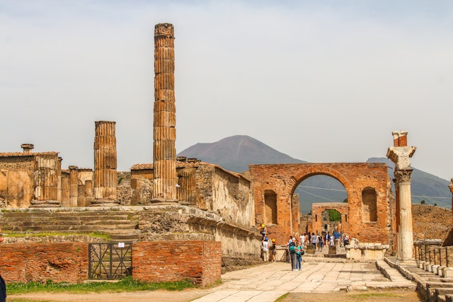 The ruined city of Pompeii. People walk around the ruins of the former city, which was destroyed by Mt Vesuvius. The volcano is visible in the background of the image.