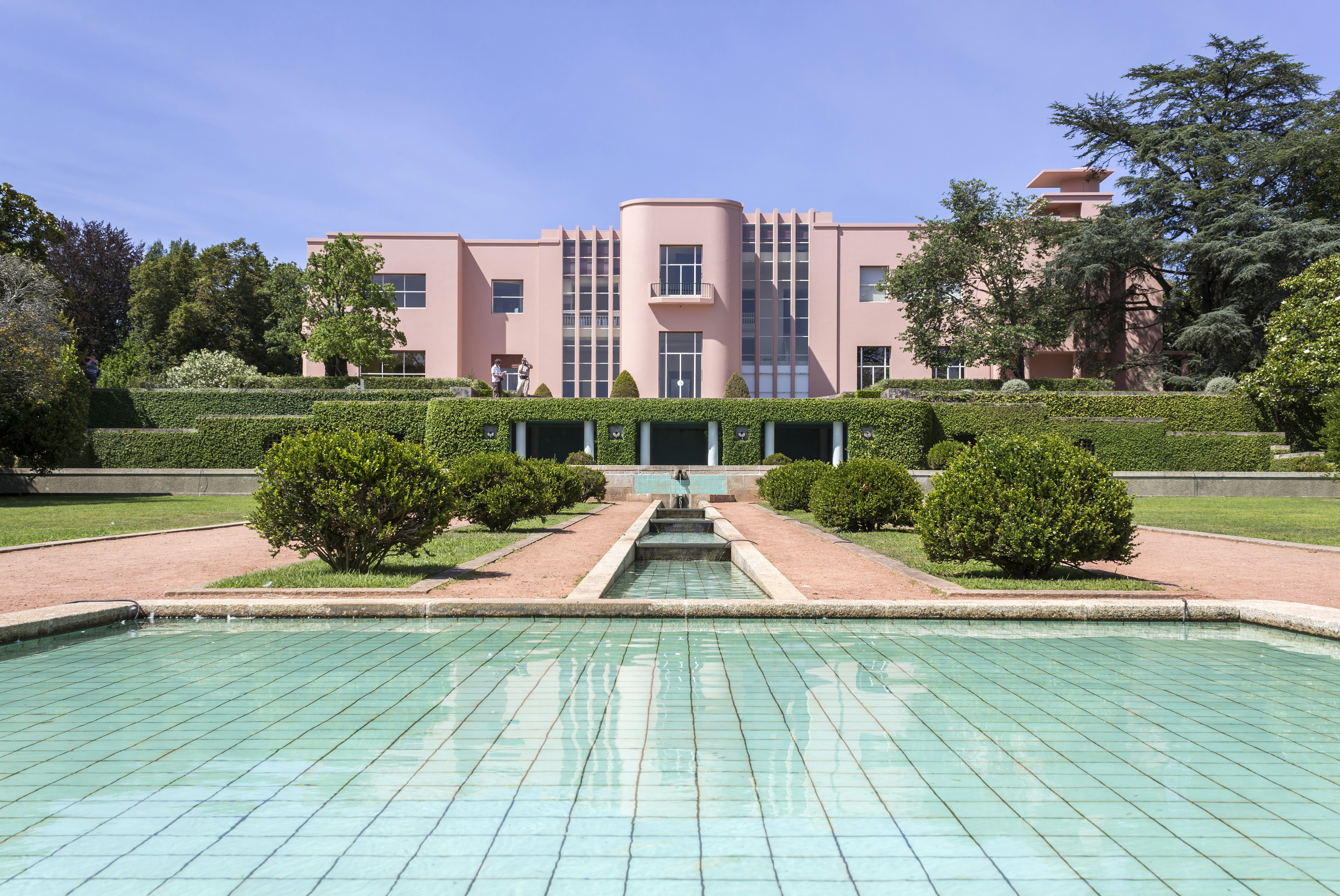 An art deco-style house in pale pink stands in manicured gardens with a central rectangular pool as a water feature.