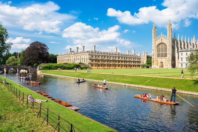 People on small flat-bottomed boats travel along a river lined by fields with large grand buildings