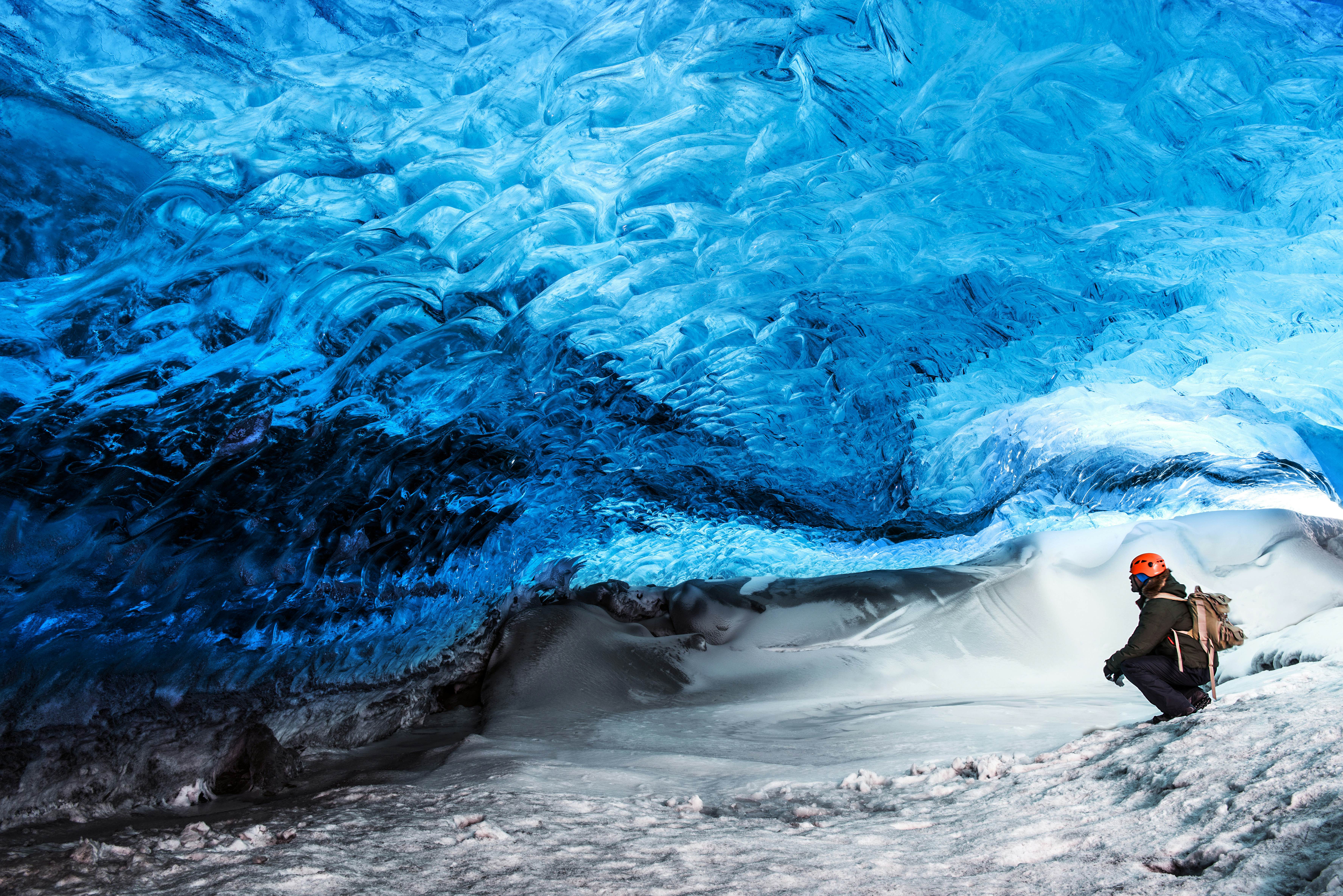 A person in safety gear squats down low inside a huge pale blue ice cave