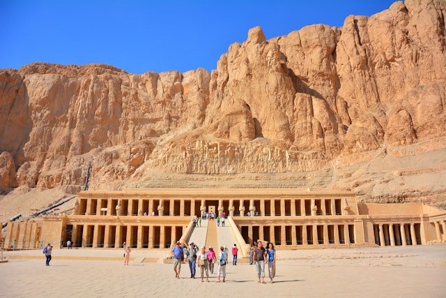 People wander in front of a vast columned temple built into a rocky hill