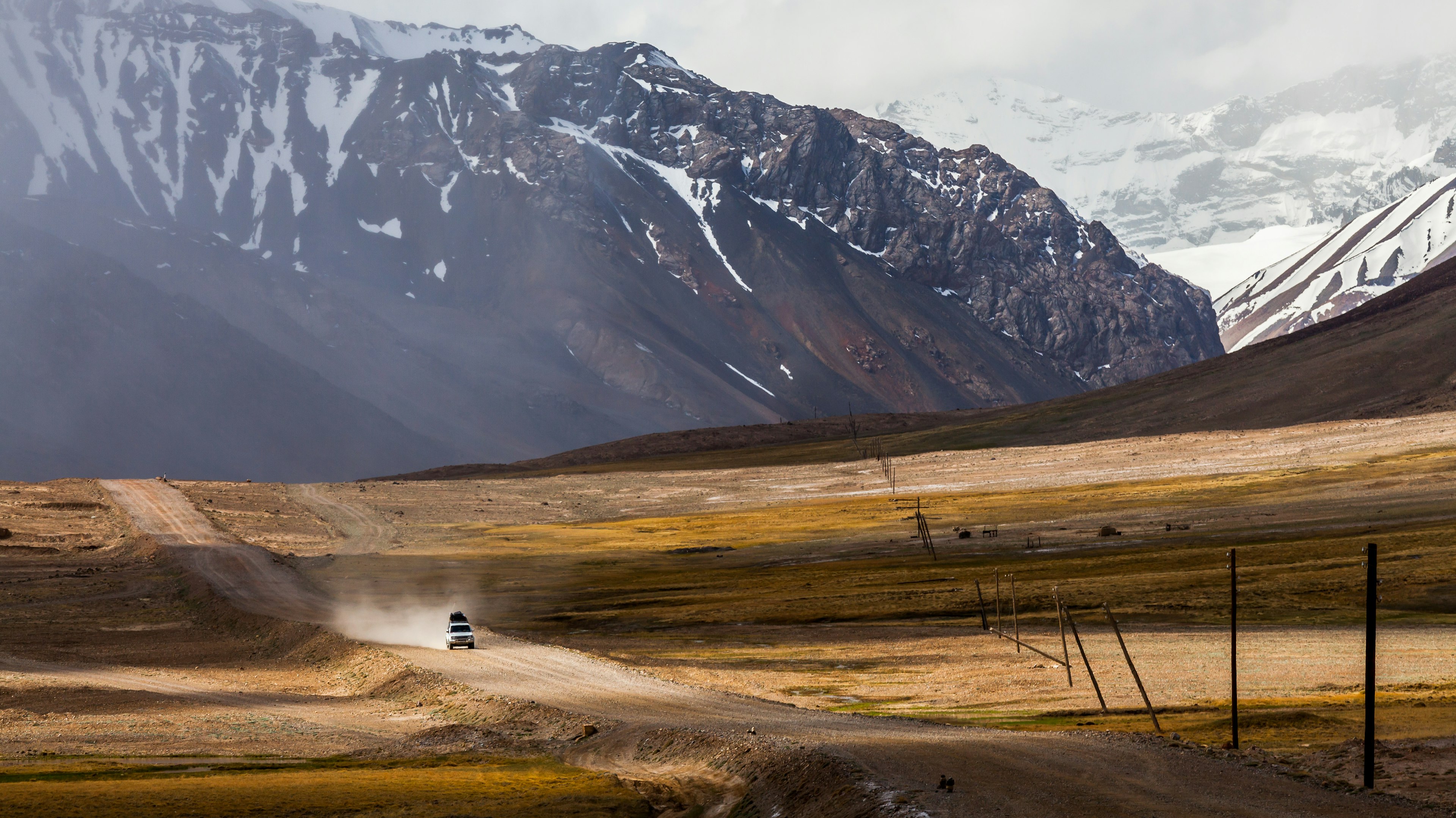 A car drives down Pamir Highway with dust in its wake, snow capped mountains in the background